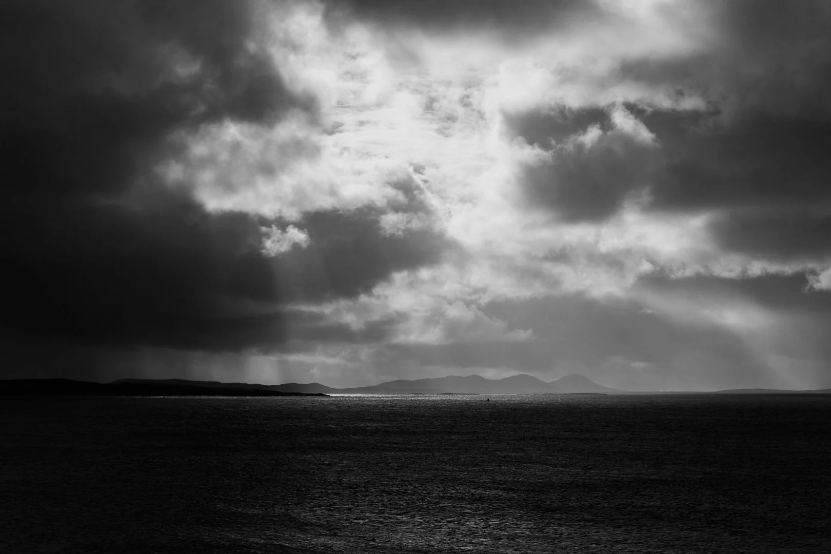 A black and white photograph of a body of water, with a distant shoreline and mountains under a cloudy sky with some sunlight breaking through.