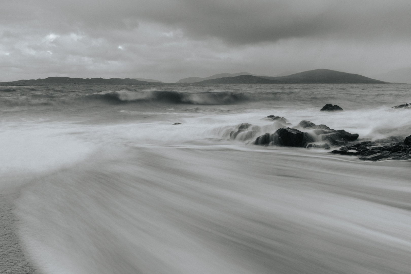 Black and white photo of ocean waves crashing onto rocky shore with distant islands and cloudy sky.