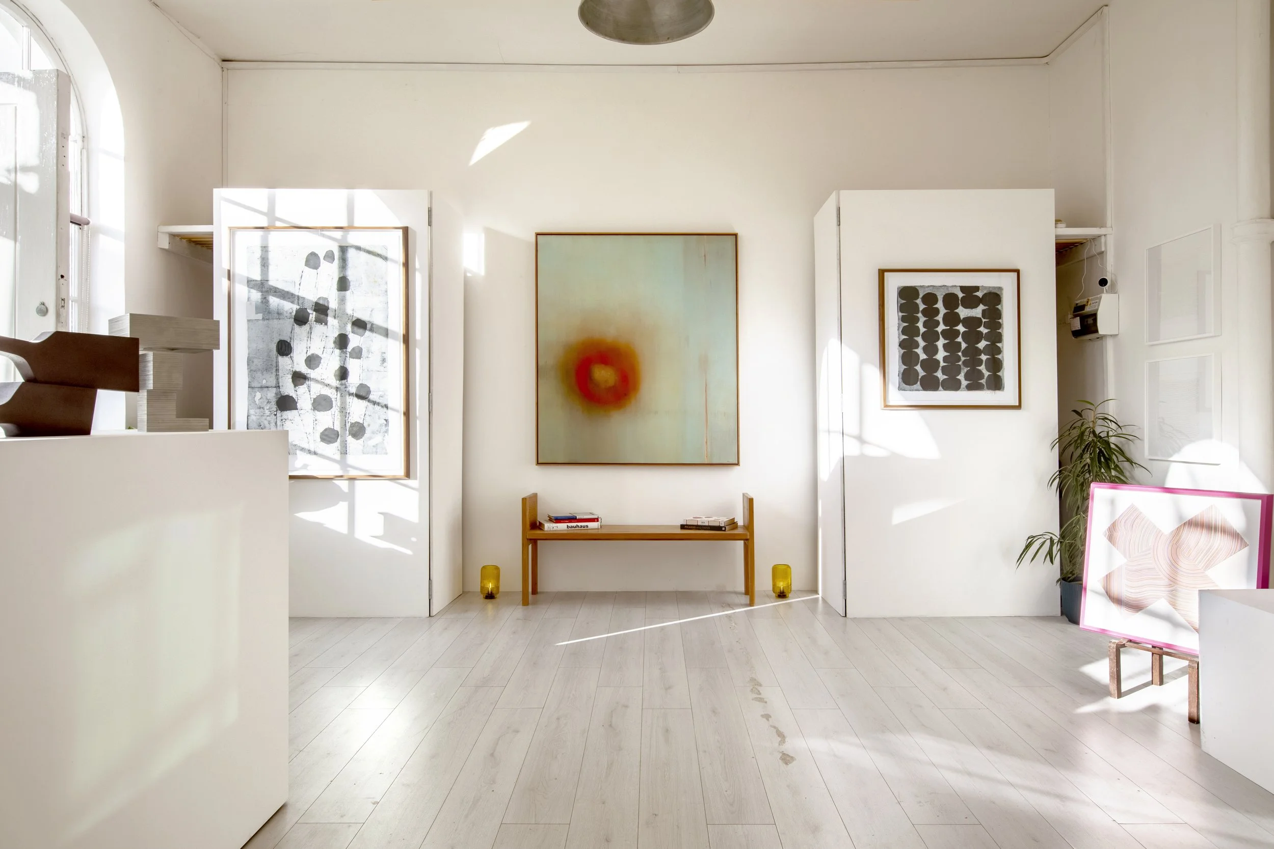 An art gallery with white walls displaying various framed artworks, a small table with wine bottles, glasses, and a sunflower centerpiece, and two grey chairs.
