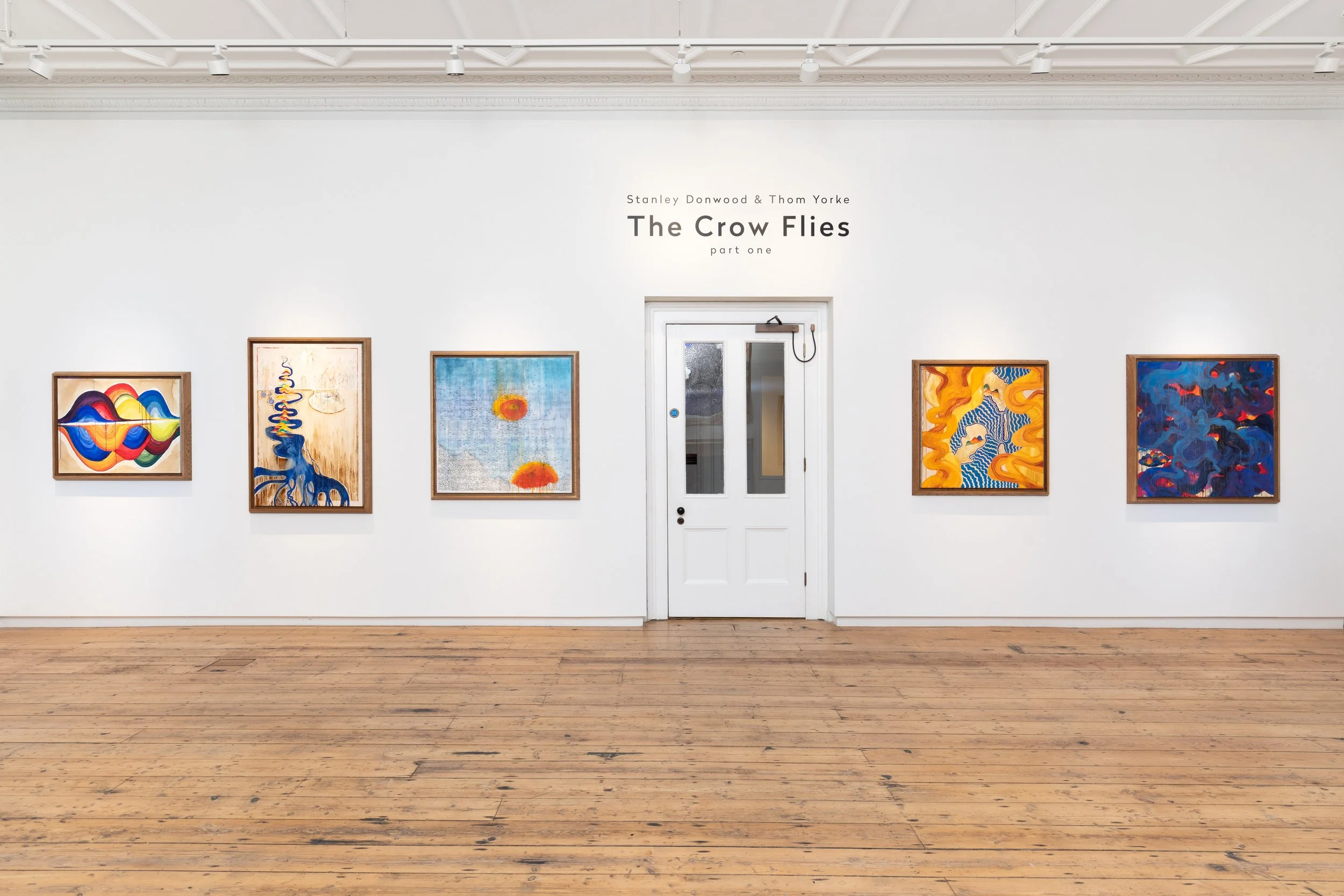 An art gallery with white walls and light wood flooring, decorated with framed abstract and landscape artwork, a potted plant, and yellow candle lanterns.