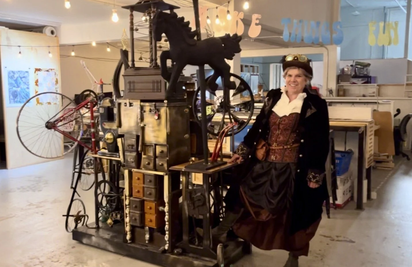 artist dressed in a steampunk outfit stands next to a fantastical machine with drawers, bicycle wheels, and a carousel horse