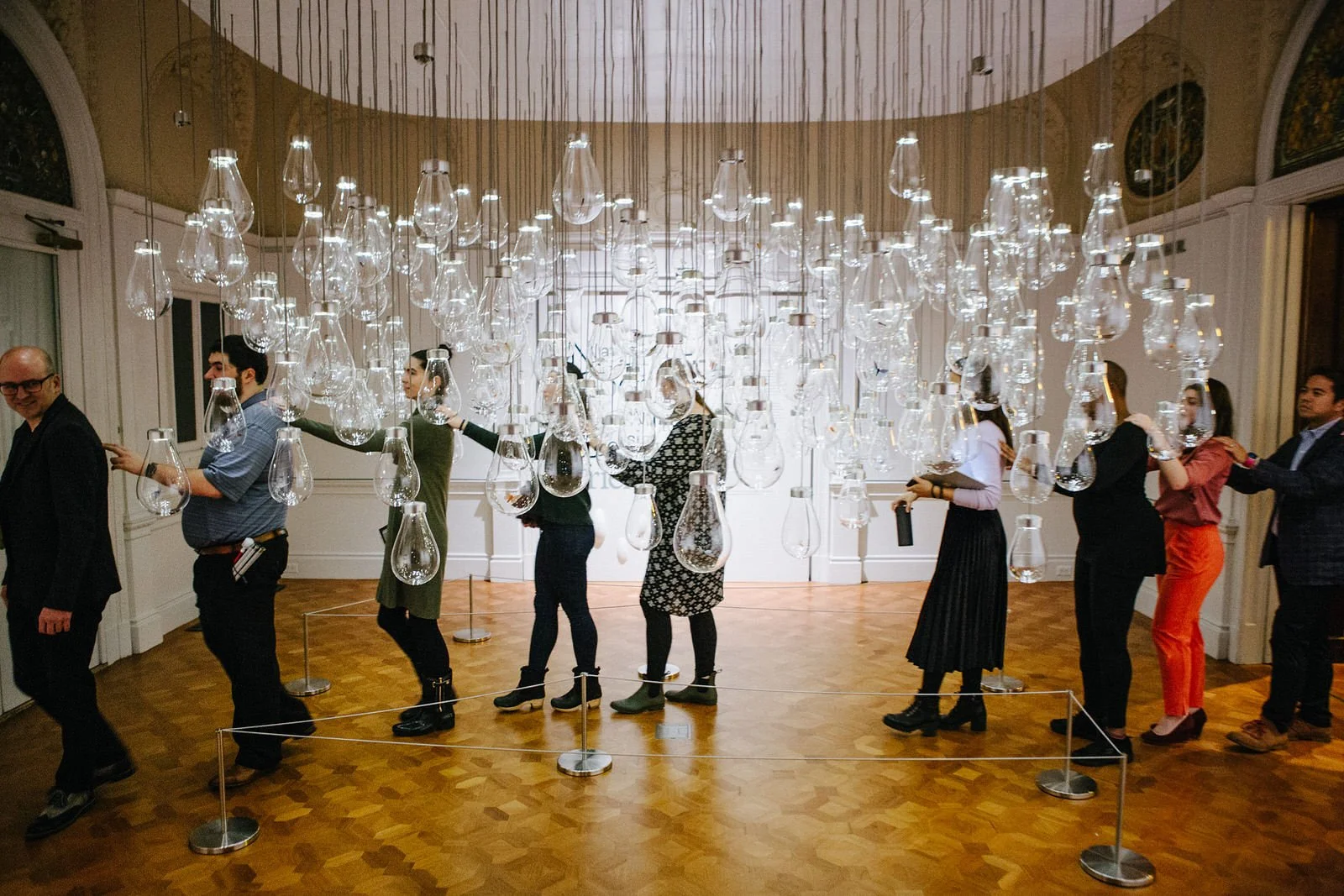 A line of people walking through a gallery at Cooper Hewitt, Smithsonian Design Museum, behind a hanging sculpture of many clear lightbulbs hung in a cluster