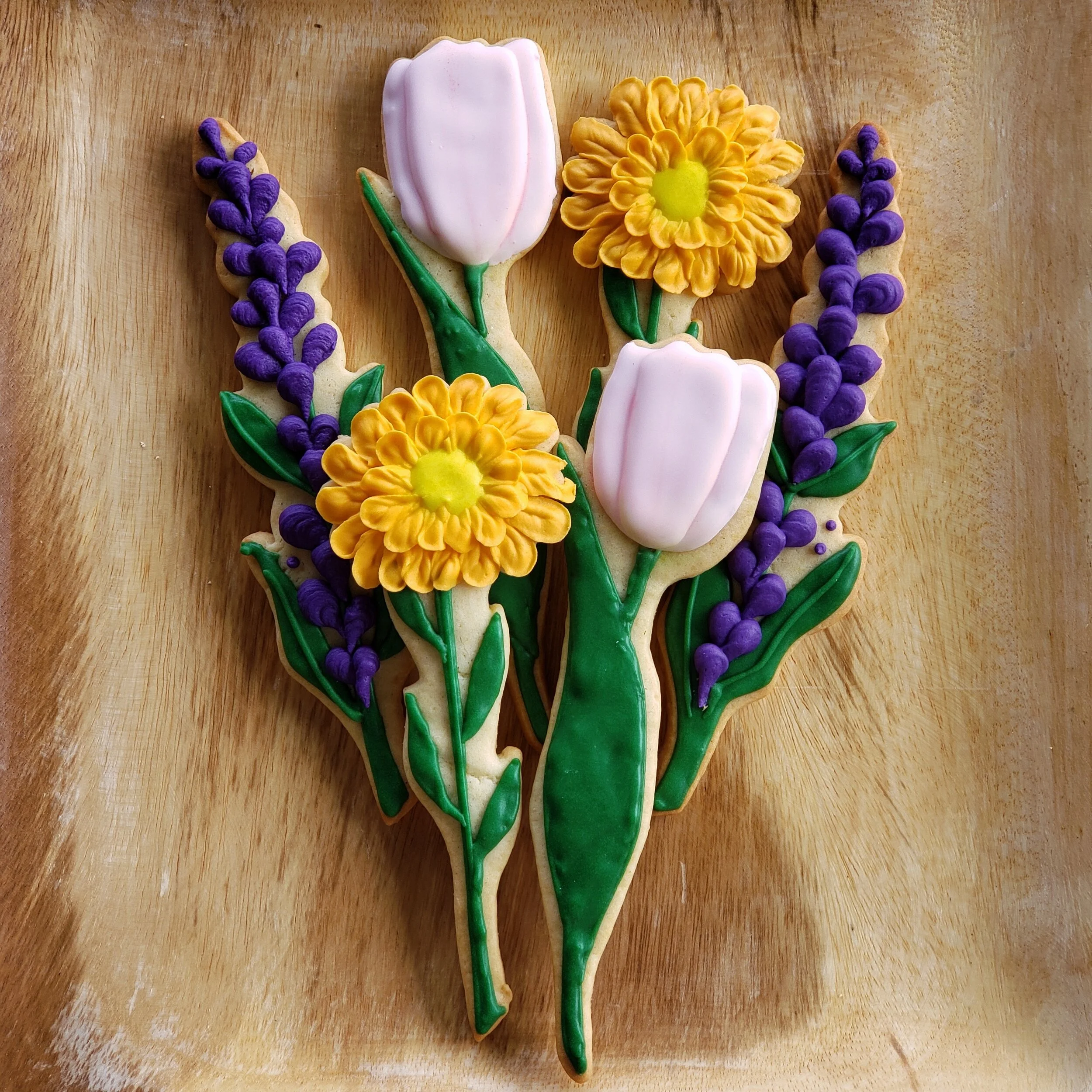 Decorative cookies shaped like flowers, including pink and white tulips, yellow daisies, and purple lavender, on a wooden surface.