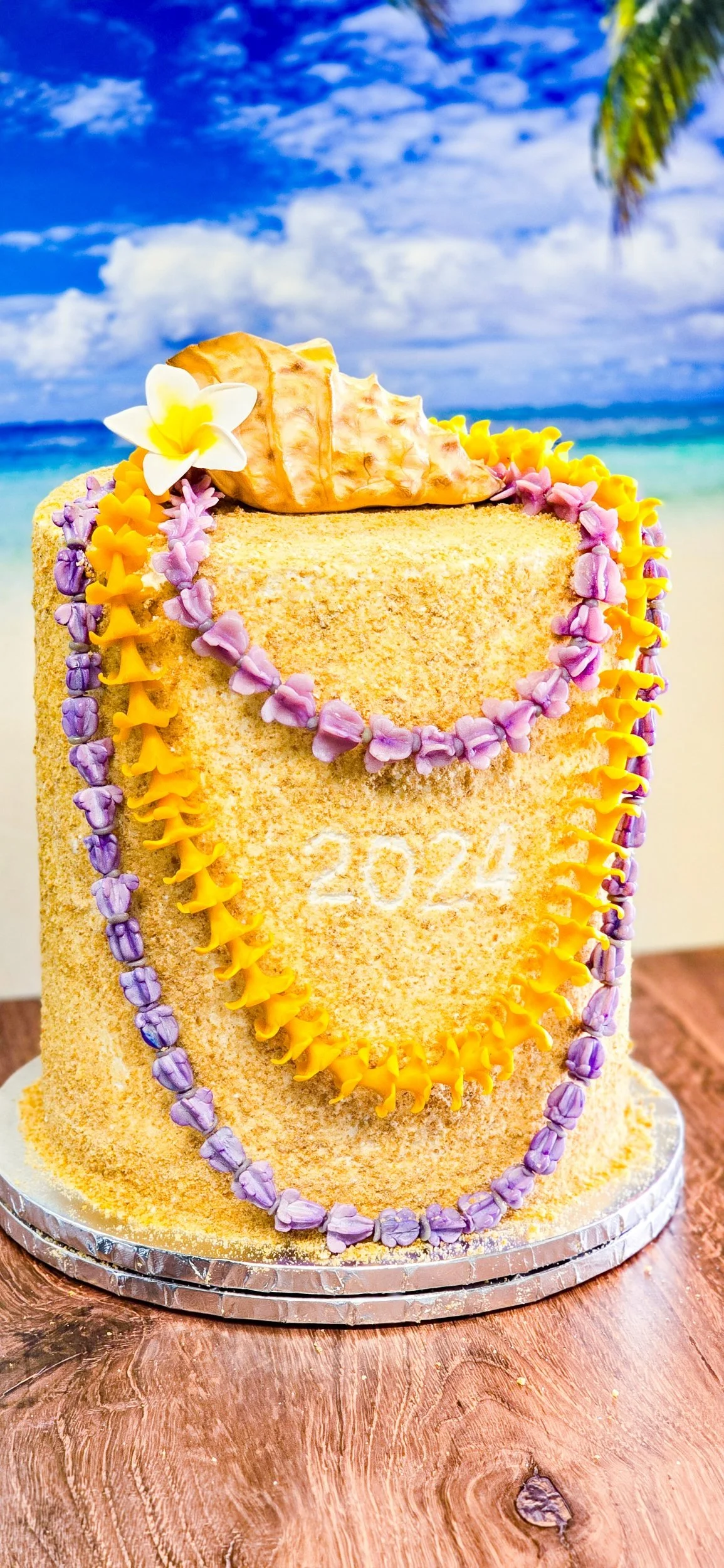 A tropical-themed birthday cake decorated with flower-shaped icing in purple and yellow, with a conch shell and a white flower on top. The cake has the year 2024 written on it and is set against a beach background with sand, ocean, and sky.