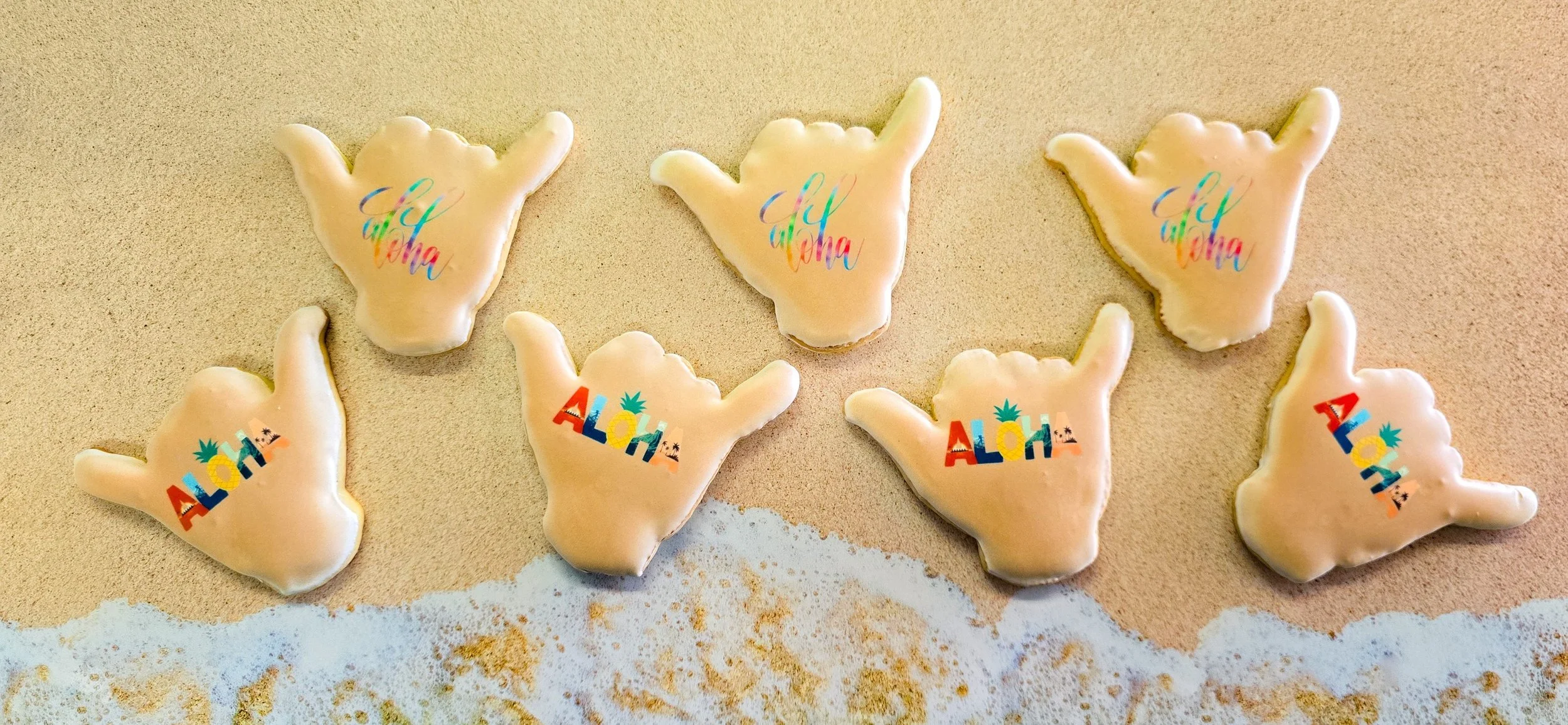 Seven cookies in the shape of hand signs, decorated with beach-themed words: four say 'Aloha' and three say 'Aloha' with colorful tropical designs, set on a sandy surface.
