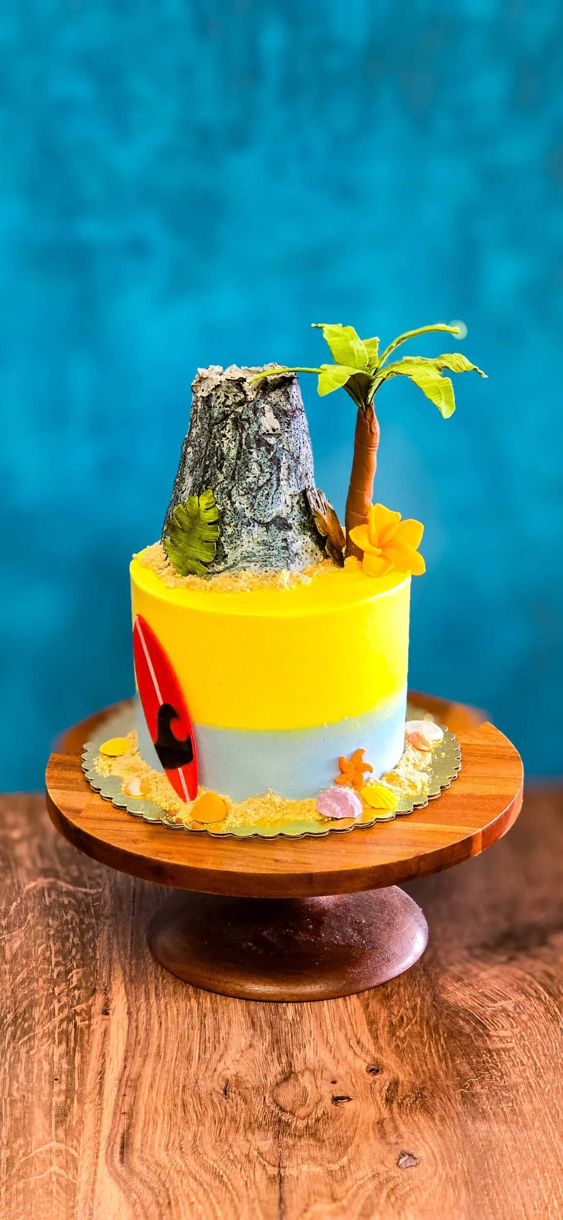 A beach-themed cake with a volcano, jungle tree, and surfboard decorations on top, placed on a wooden cake stand.