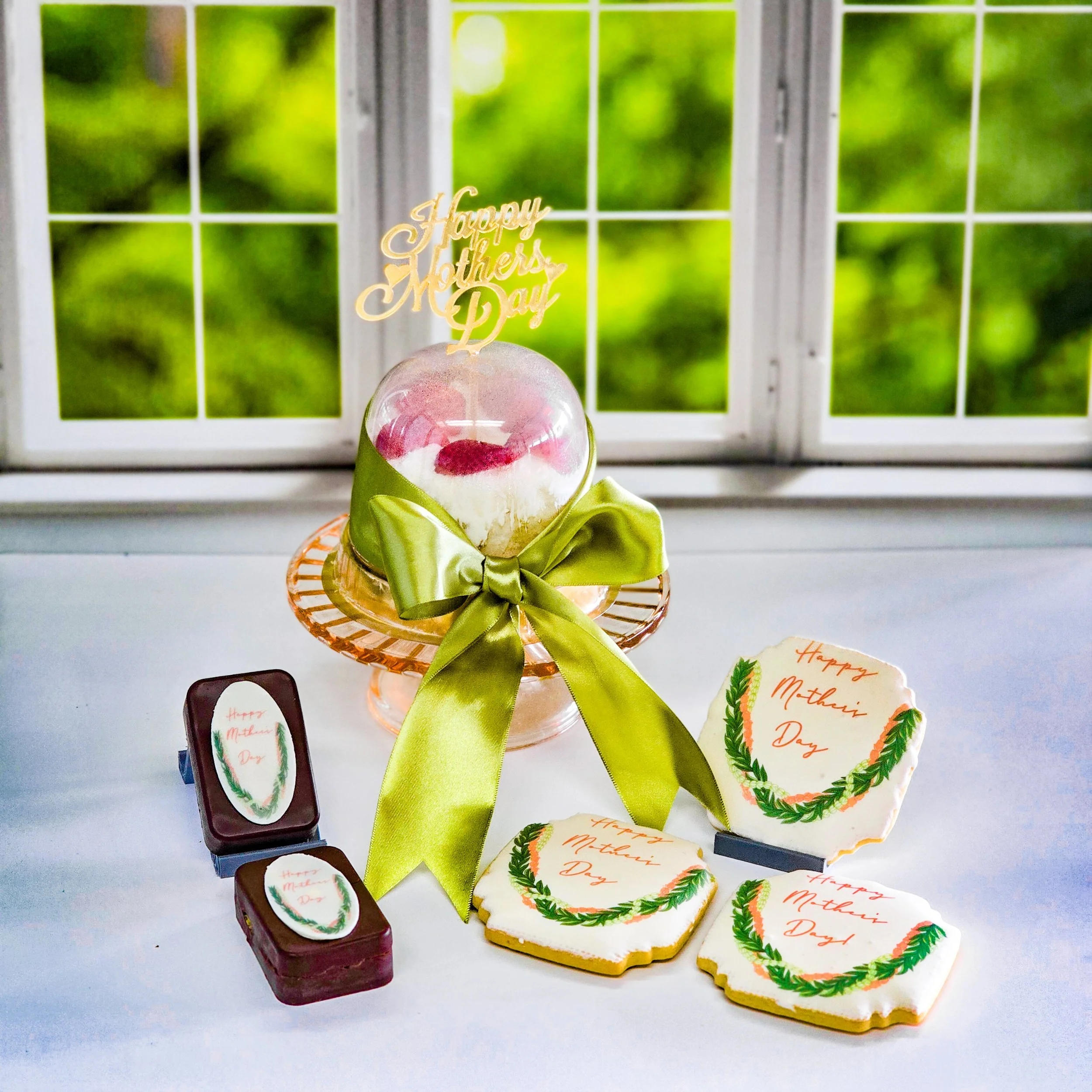 Mother's Day celebration with decorated cookies, chocolates, and a cake topped with a "Happy Mother's Day" sign and a green ribbon, set on a white surface near a window with a green outdoor view.