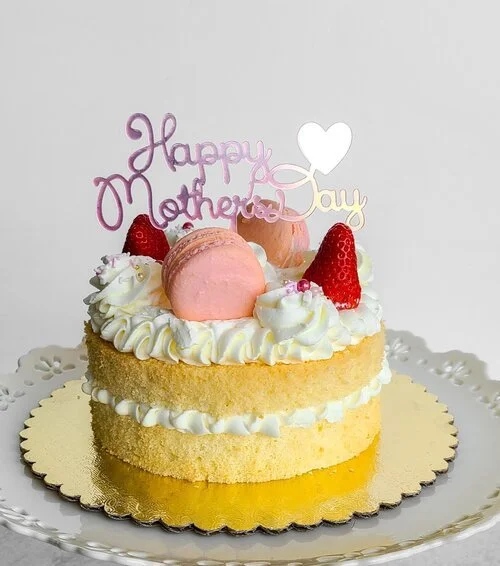 A Mother's Day cake with two layers of yellow sponge, decorated with whipped cream, strawberries, and pink macarons. It has a topper reading 'Happy Mother Day' with a heart, placed on a gold cake board.