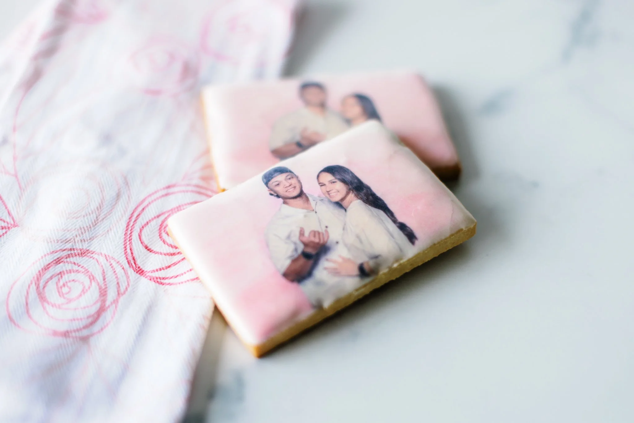 Two decorated cookies with printed images of a couple, placed on a white surface with a pink cloth featuring red spiral designs.