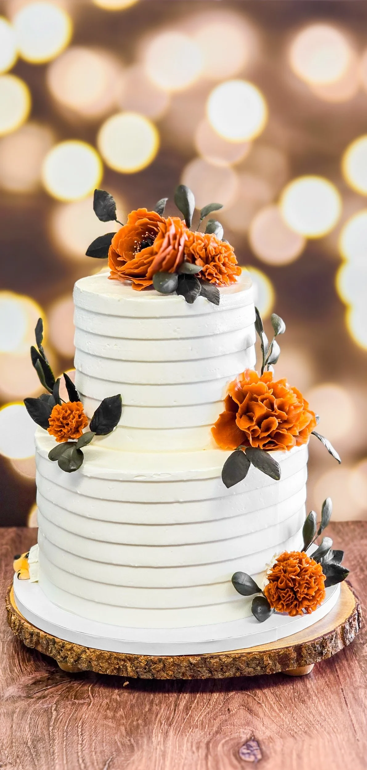 A two-tiered white frosted cake decorated with orange flowers and green leaves, placed on a wooden cake stand with a blurred golden bokeh background.
