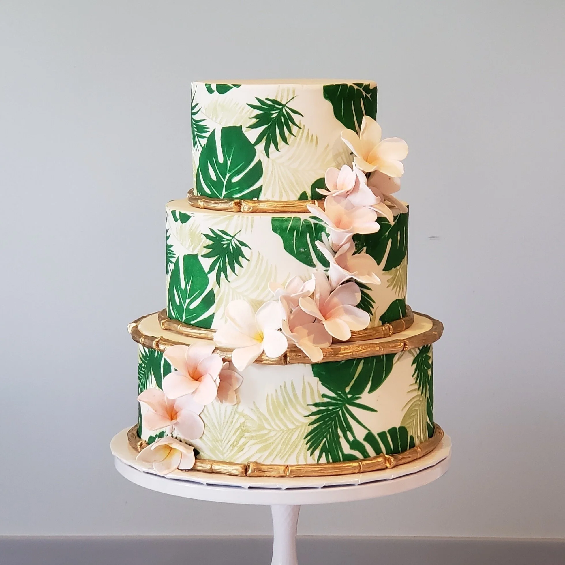 Three-tier tropical-themed cake with green leaf patterns, pink flowers, and gold accents on a white cake stand - custom wedding cake at Short N Sweet Bakery and Cafe in Hilo, HI.