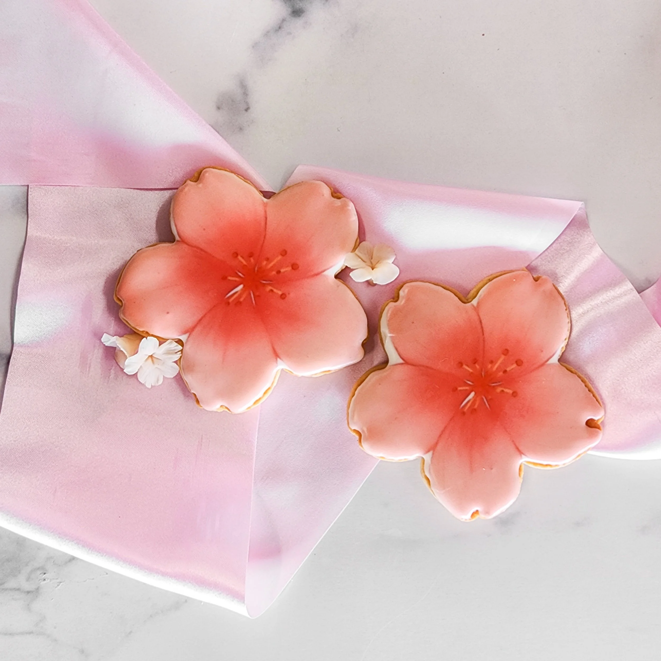 Decorative cookies shaped like pink cherry blossoms with small white flowers on a pink satin cloth.