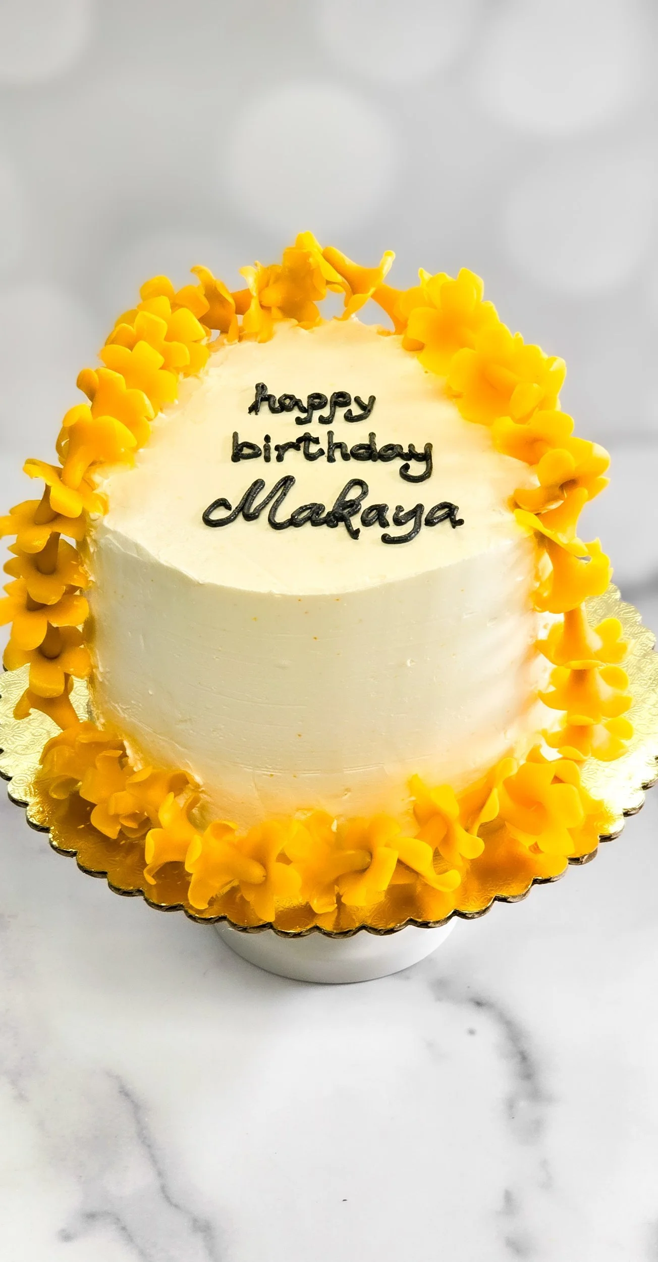 A round white birthday cake decorated with yellow flower-shaped frosting around the top edge, with the words "happy birthday Makaya" written in black icing on top, placed on a marble surface.