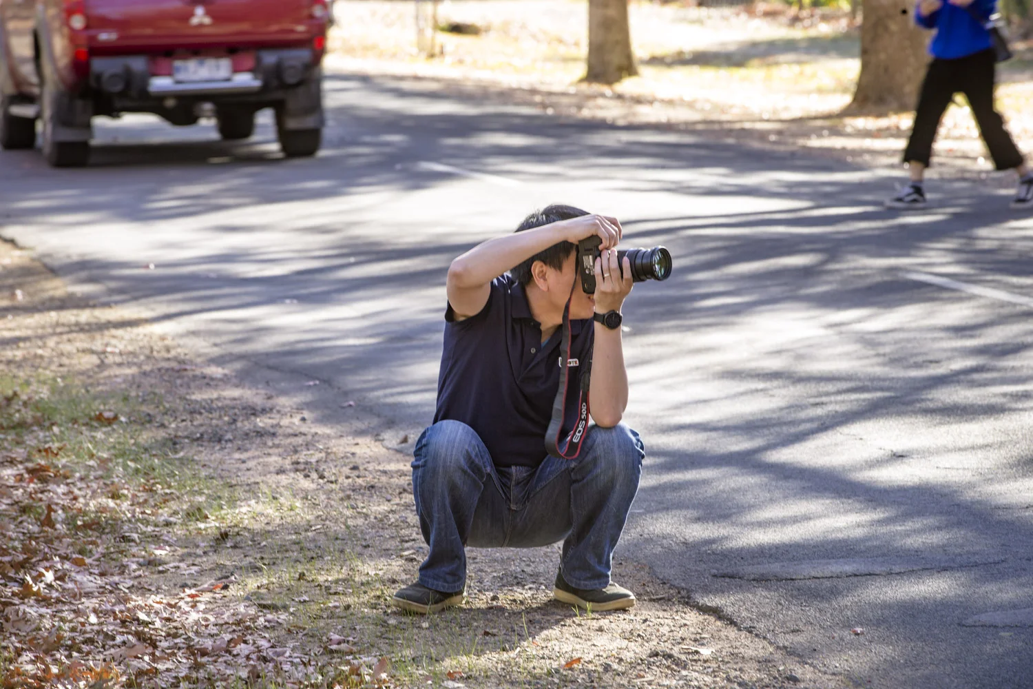 A photographer squats on the roads edge taking images of another on the road, focused on the image, forcing cars to move around him.