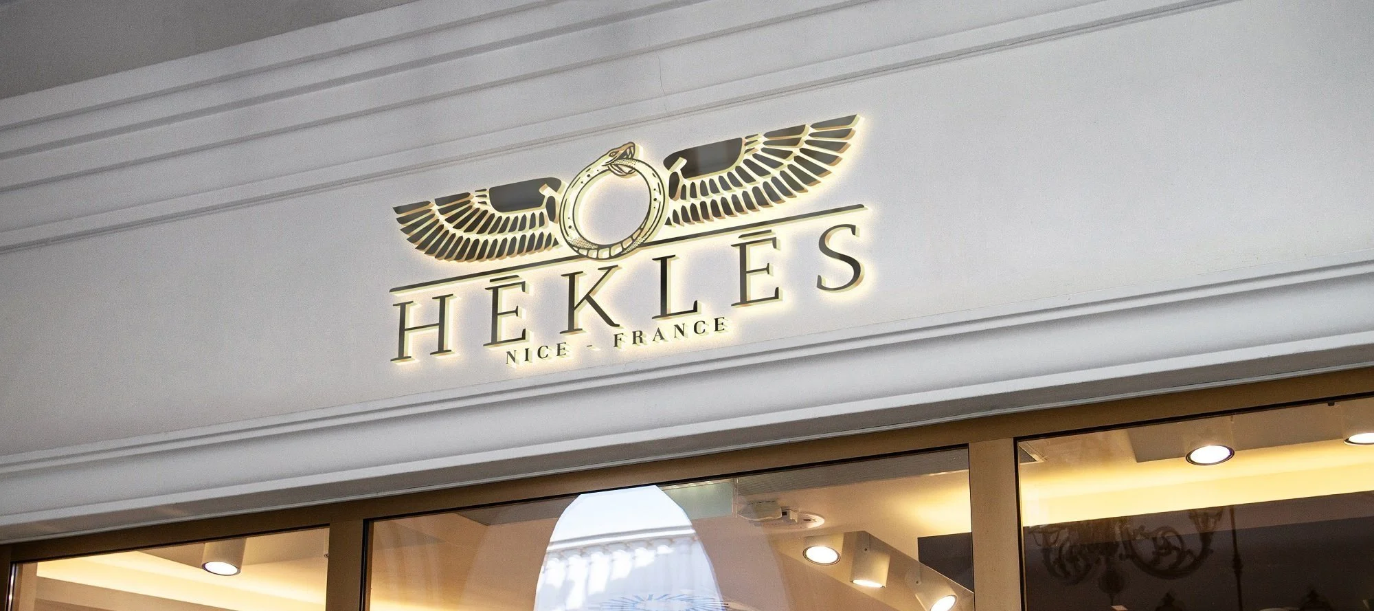 HÉKLÈS winged-ouroboros logo, back-lit in gold on a shop facade.