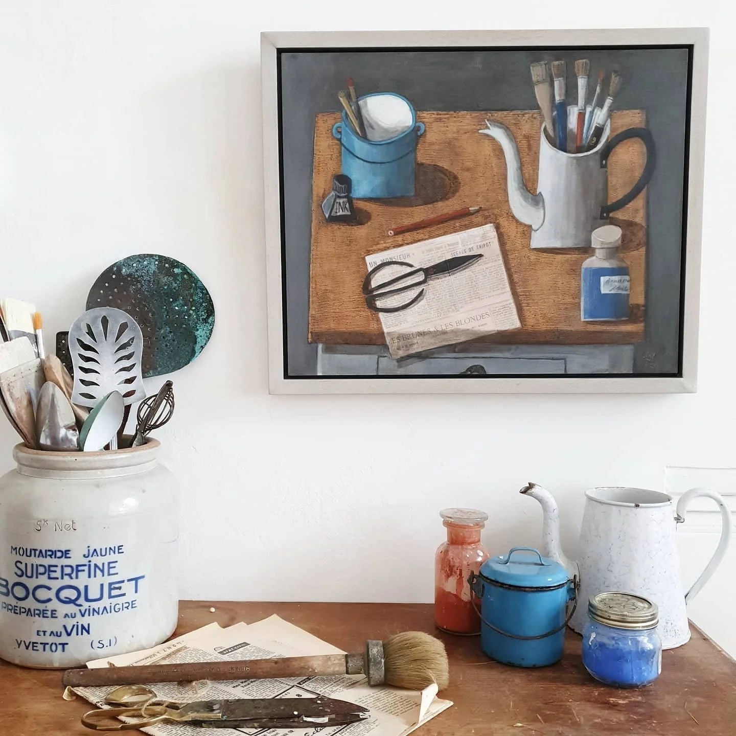 Artist studio table with brushes, pigment jars and a framed still life painting on the wall.