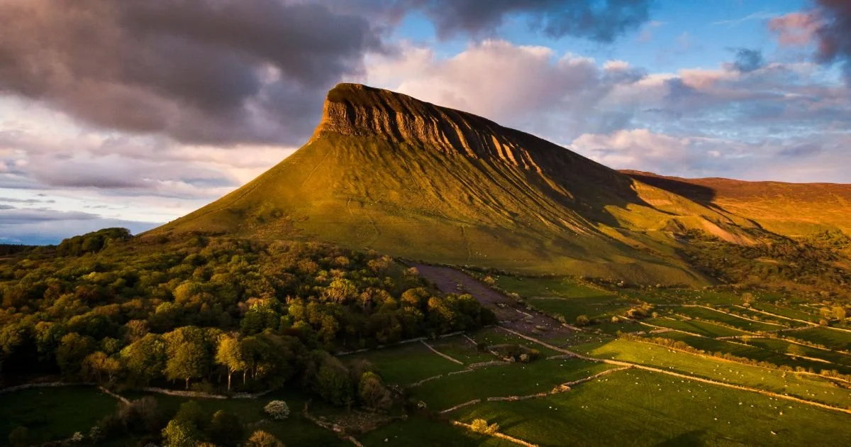 the green majestic heights of Ben Bulben Mountain in Sligo, west of Ireland. Green fields lie at the bottom of the mountain, delineated by stone walls.