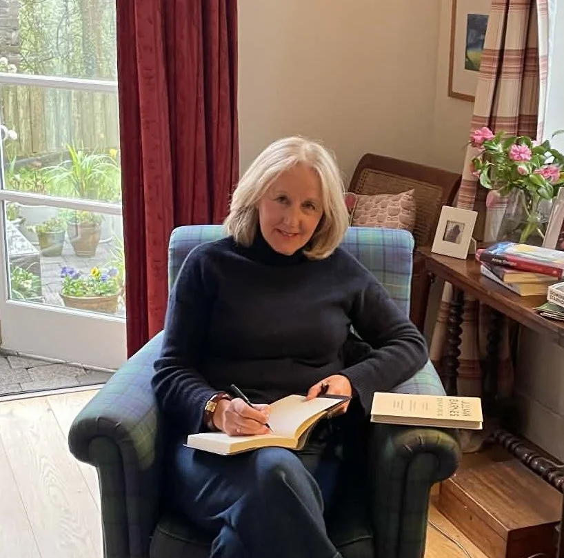 Writer Marion Garvey sitting in an armchair, writing in her notebook, with a book beside her
