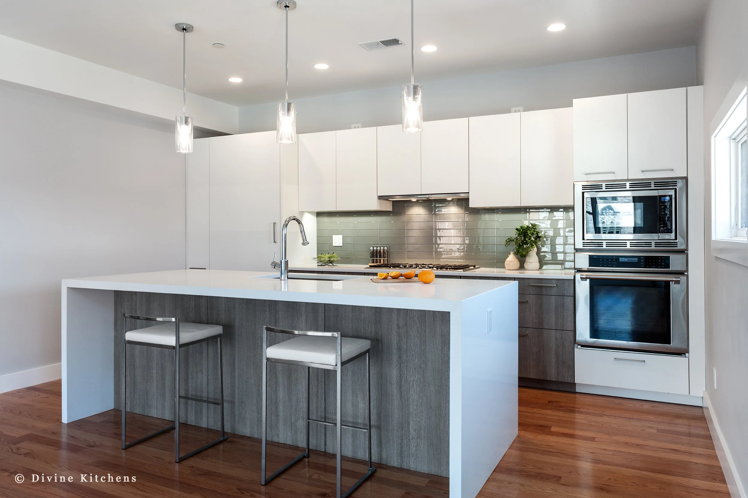 Modern kitchen with matt white, flat-panel cabinets and cambria countertops with a waterfall edge on island, green subway tile backsplash, wall oven.