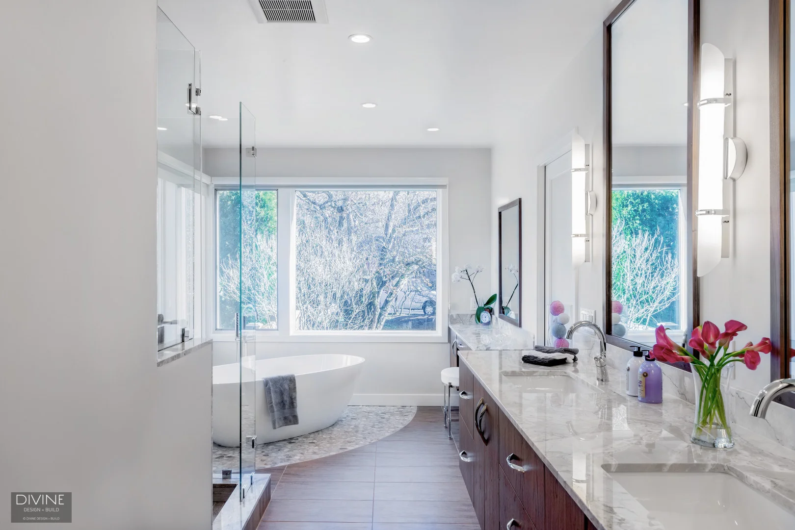 Modern bathroom with white stand alone tub, combination porcelain and natural stone floor tiles. Curbed shower with glass partition. Floating his and her cherry wood vanities, and marble countertops.