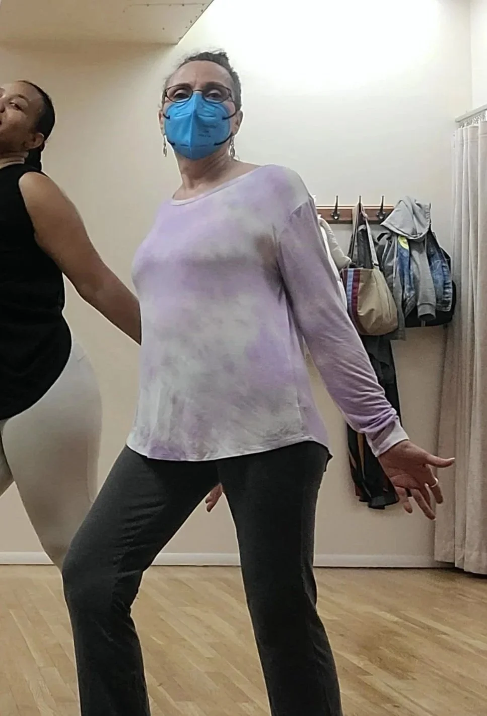 An older Latina woman with glasses in a face mask striking a dance pose in a dance studio