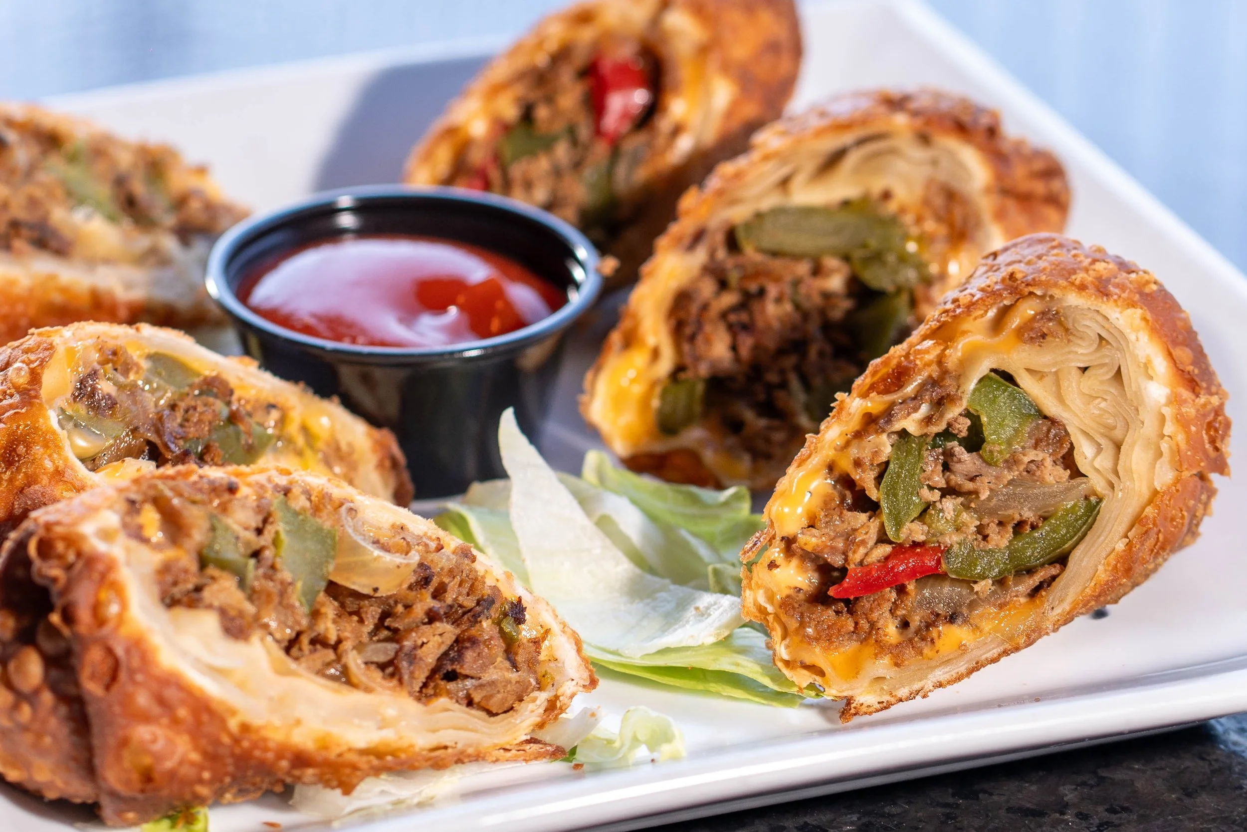 Fried egg rolls with beef, vegetables, and cheese, served with a side of red dipping sauce on a white plate.