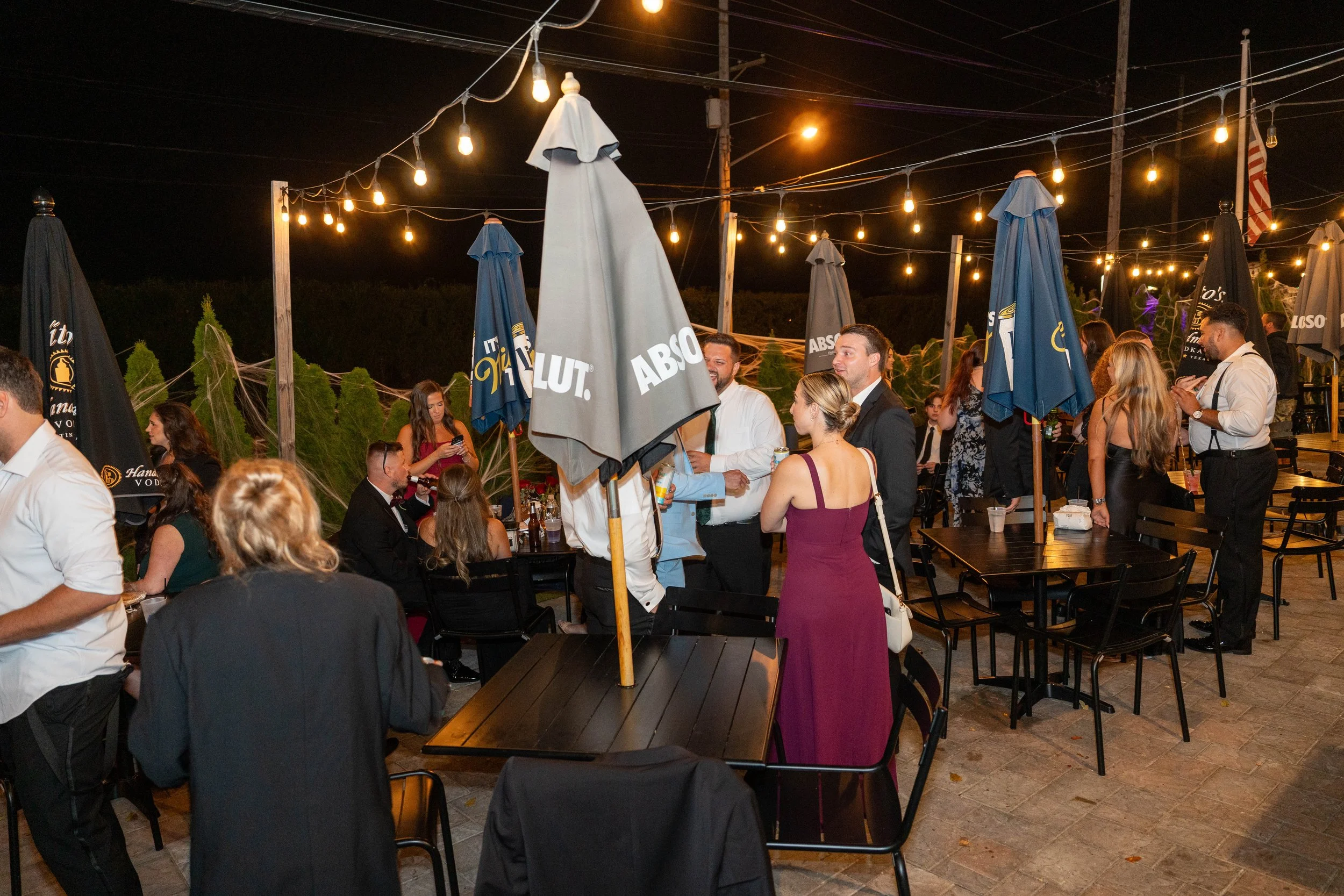 People socializing at an outdoor nighttime gathering on a patio, with string lights overhead, tables, umbrellas, and some individuals holding drinks.