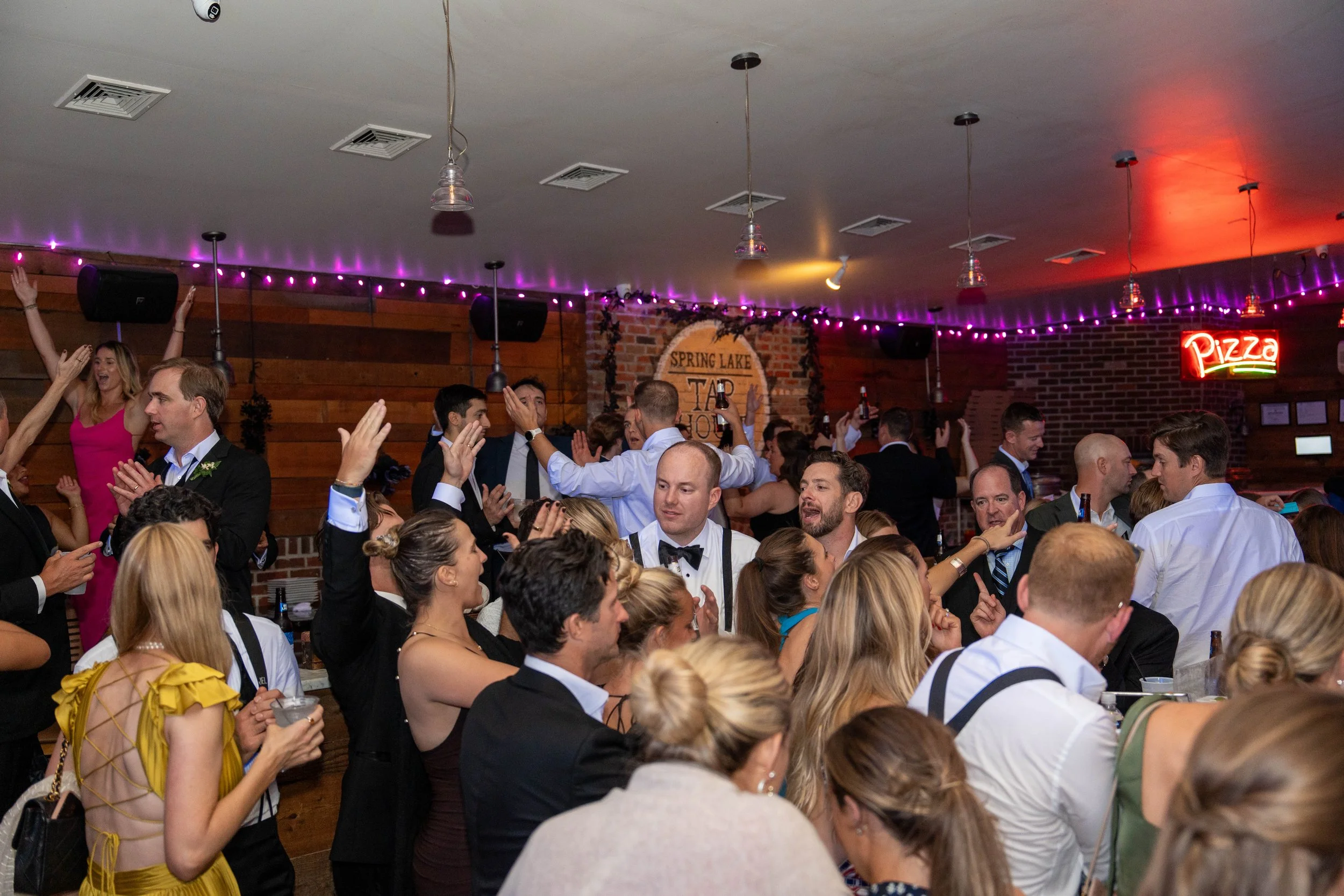 Crowd of people dancing and celebrating at a wedding reception in a decorated indoor venue with purple string lights, a pizza sign, and a sign that reads 'Spring Lake Tap House' in the background.