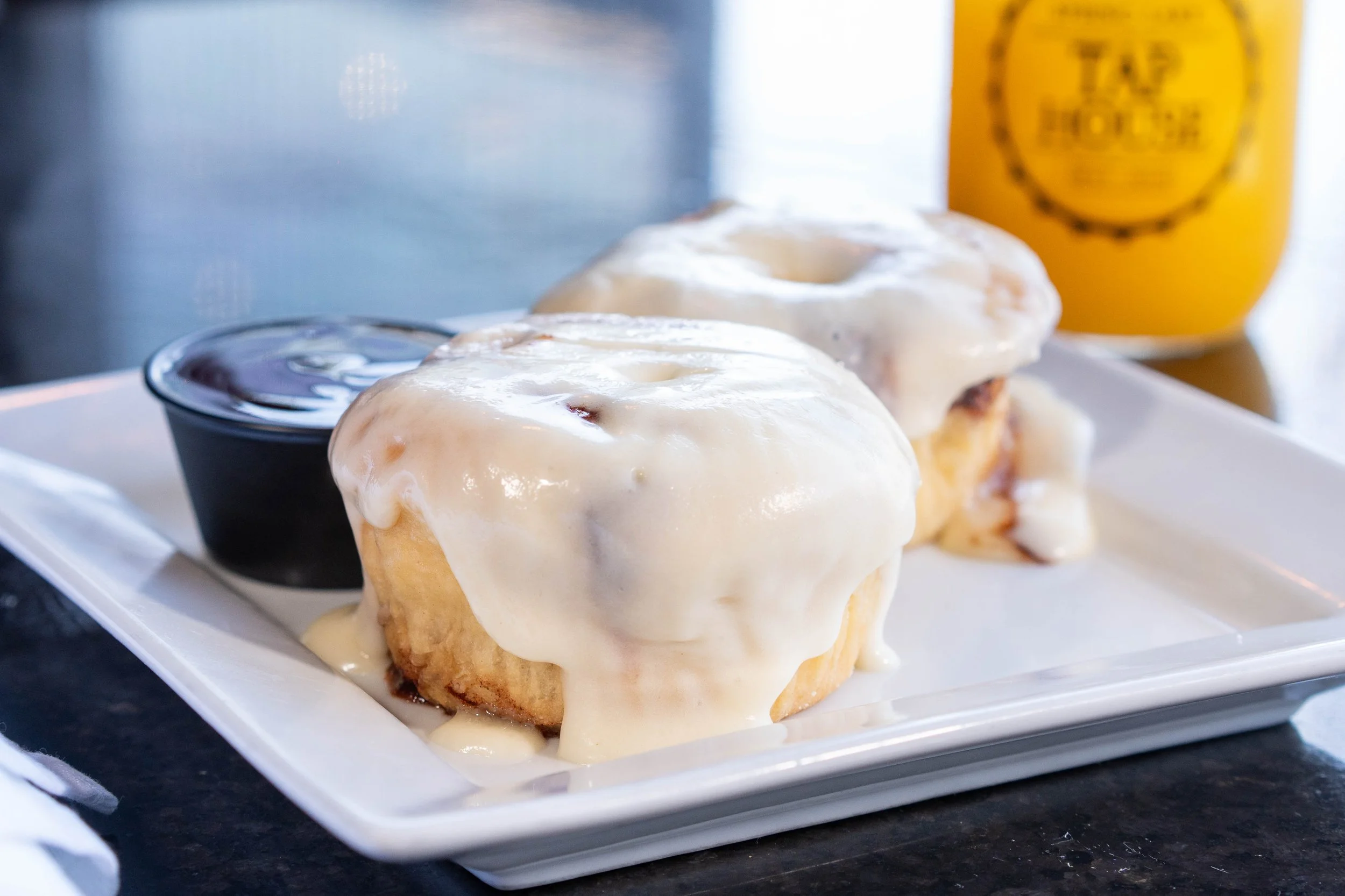 Two cinnamon rolls with white icing on a white rectangular plate, with a small cup of chocolate syrup and a glass of orange juice in the background.