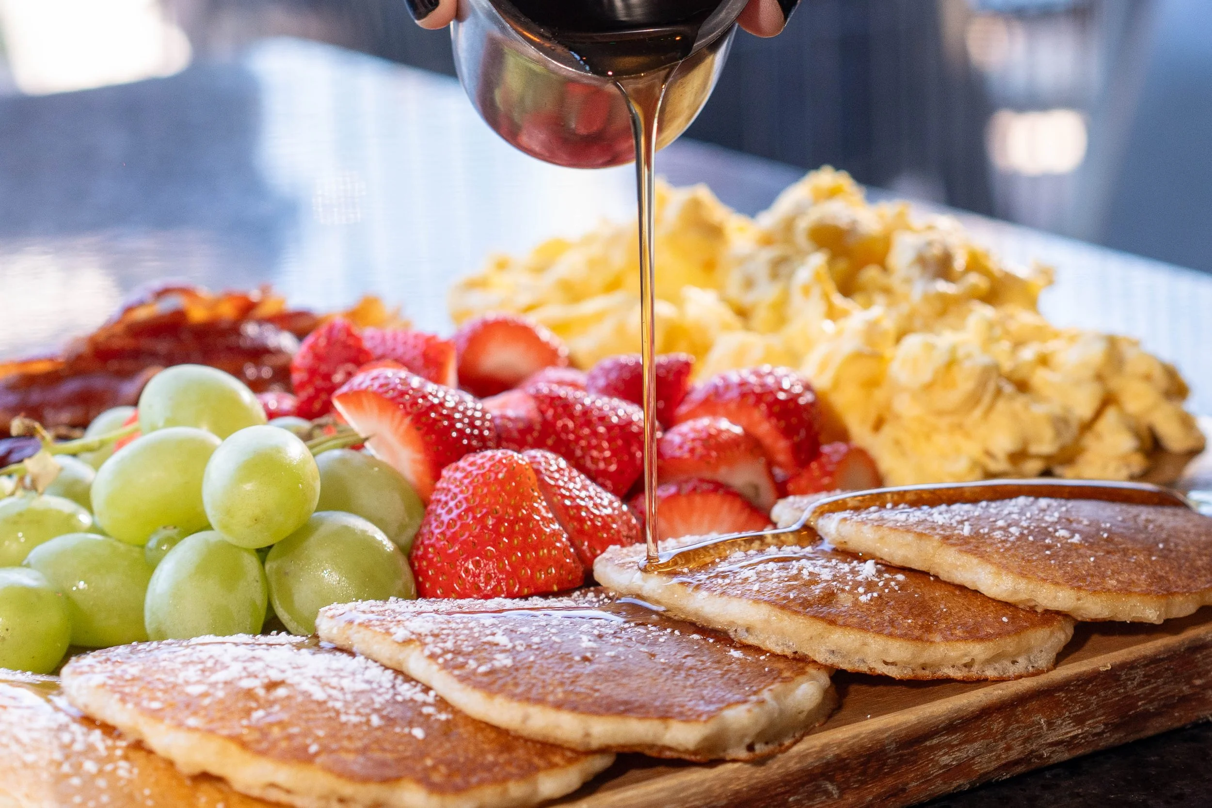 A plate of pancakes topped with powdered sugar, surrounded by green grapes, strawberries, scrambled eggs, and bacon, with syrup being poured over the pancakes.