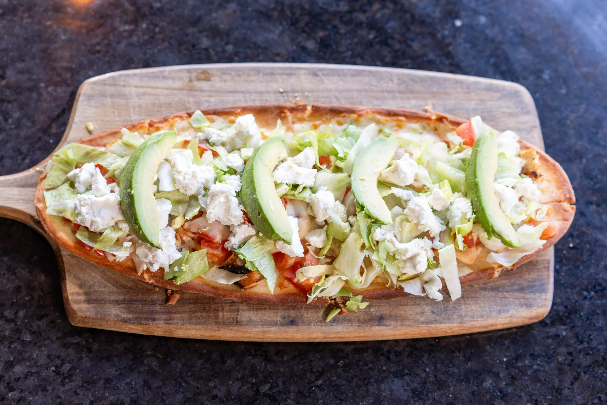Open-faced taco on wooden board with avocado slices, shredded lettuce, cheese, and tomato.