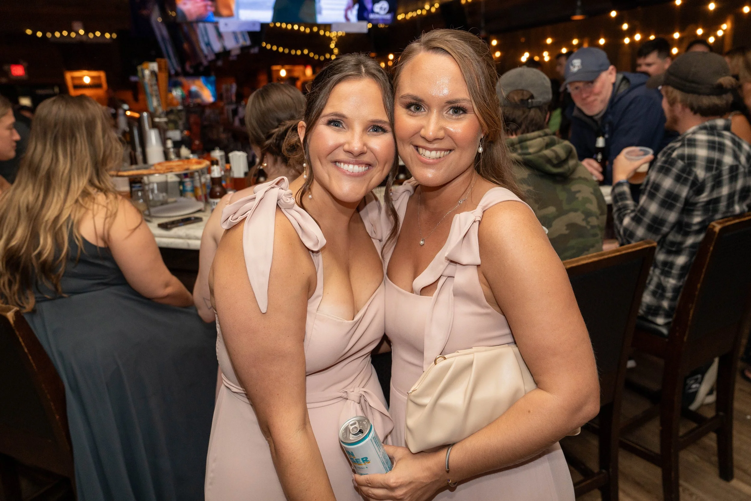 Two women smiling and posing for a photo at a lively bar or restaurant with other patrons in the background, ambient lighting, and television screens.
