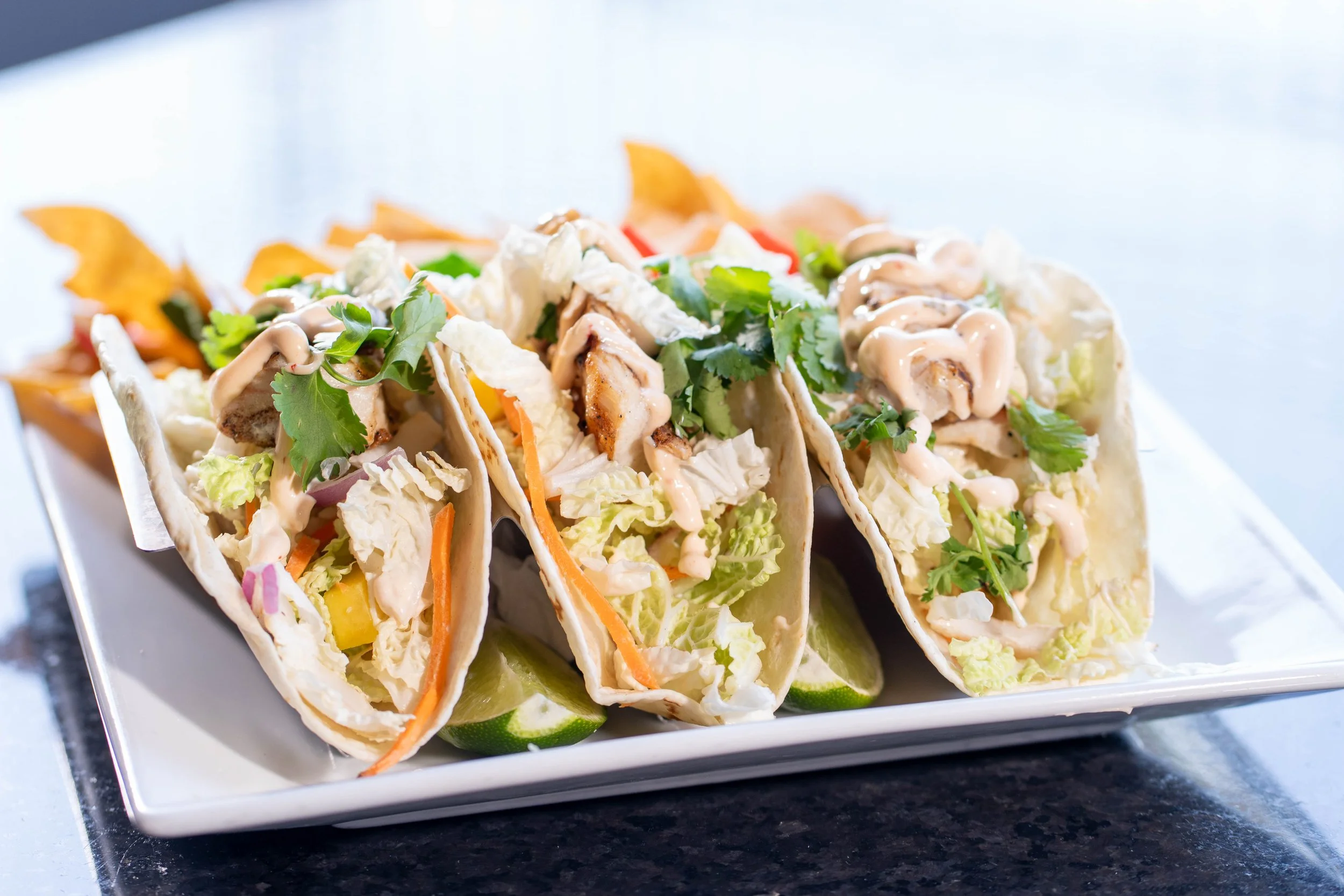 Three tacos filled with shredded lettuce, grilled chicken, sliced vegetables, cilantro, and drizzled with sauce, served on a white rectangular plate with lime wedges and tortilla chips in the background.