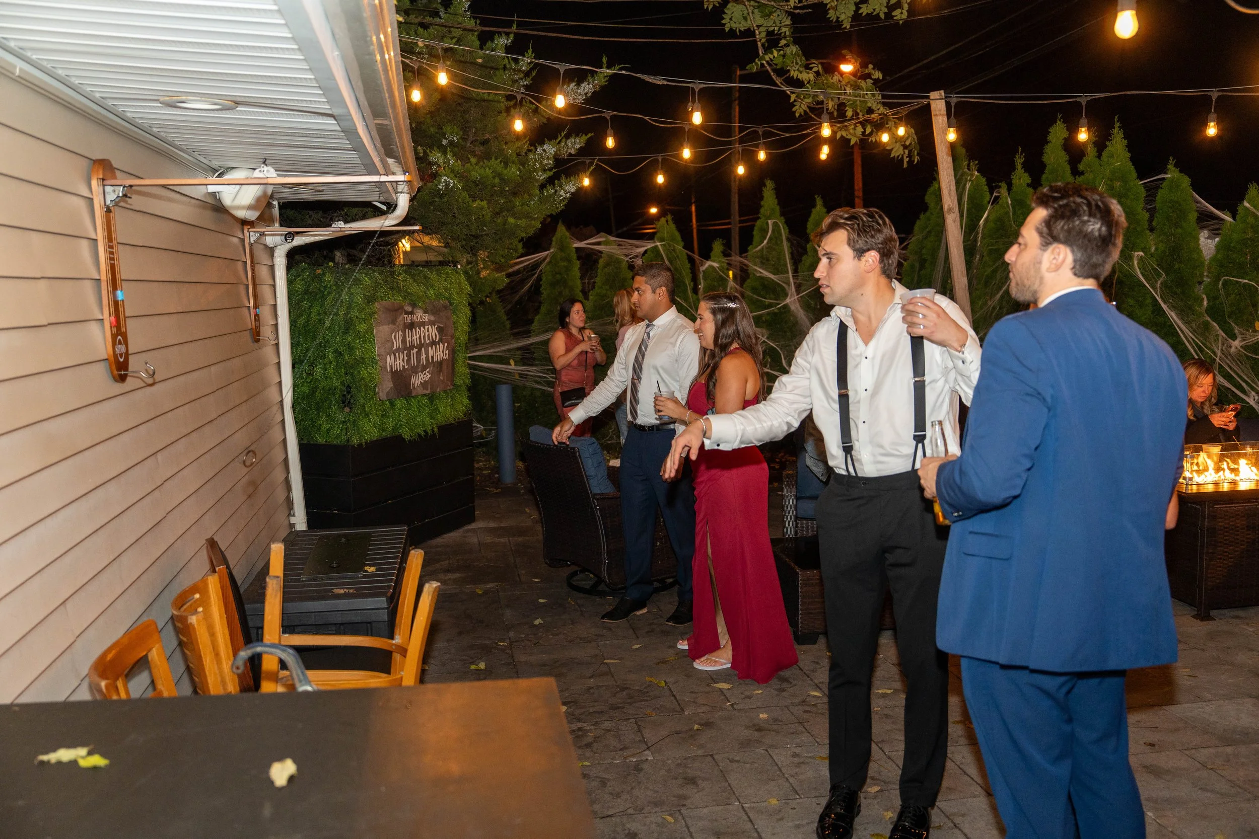 People dressed in formal attire socializing at an outdoor nighttime gathering, with string lights overhead, and decorative cobwebs in the background.