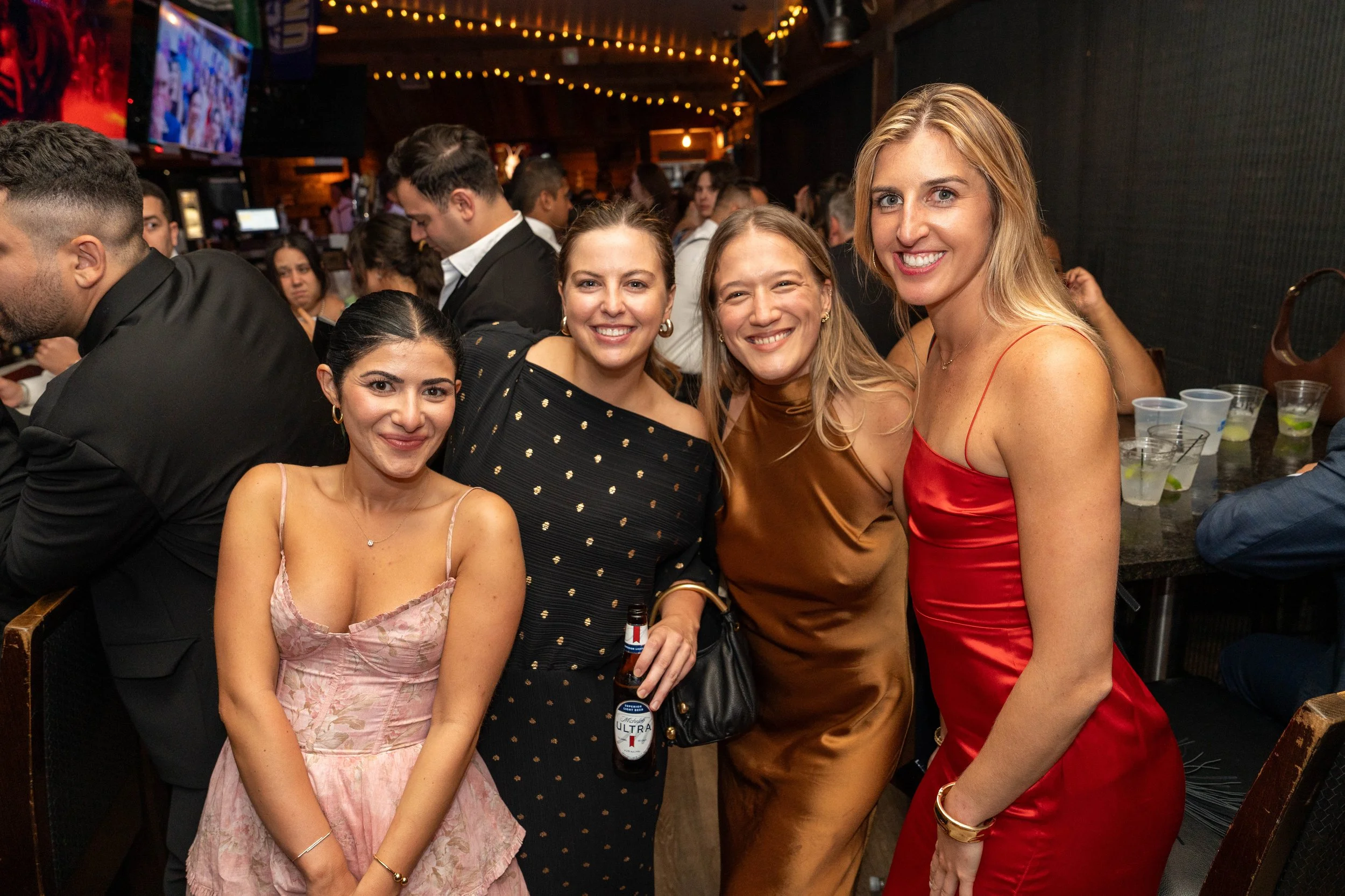 Four women smiling and posing together at a bar or nightclub, with other people in the background.