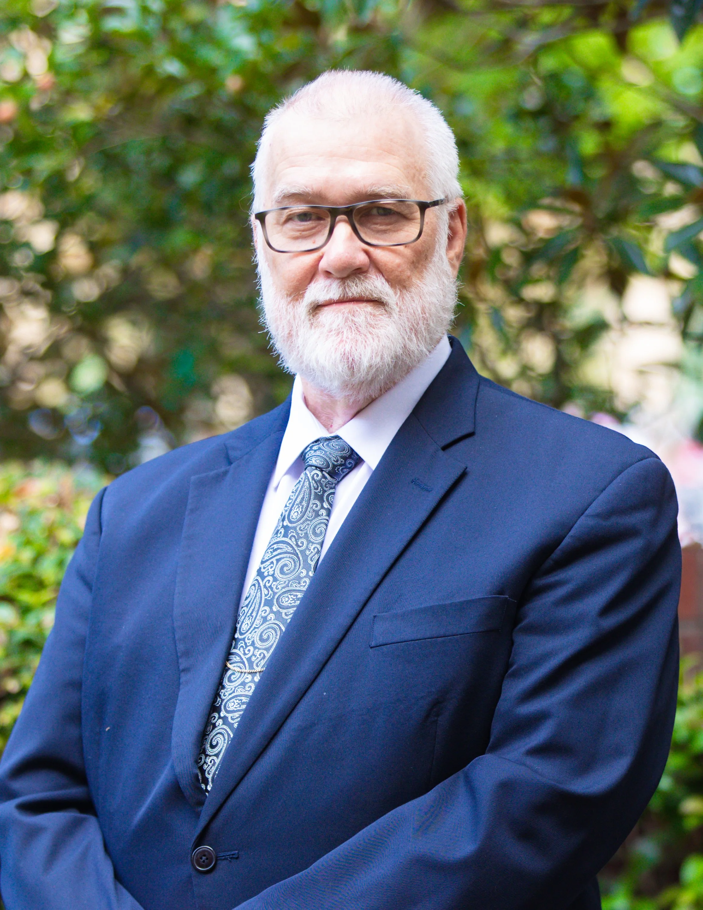 An older man with white hair, beard, and glasses wearing a dark blue suit, white shirt, and patterned tie, standing outdoors with trees and greenery in the background.