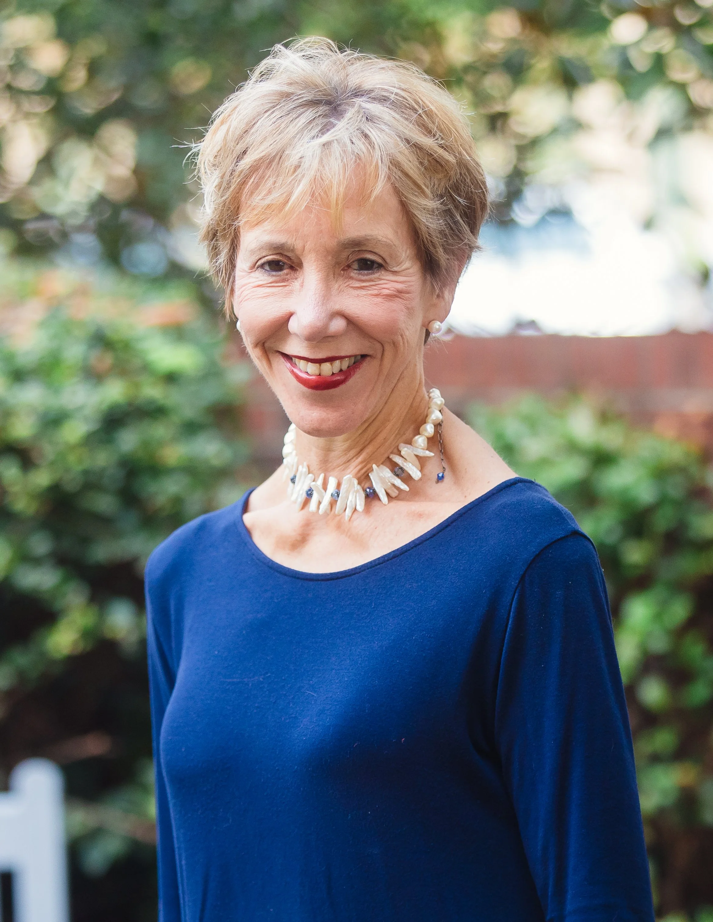 A smiling woman with short blonde hair, wearing a blue top, pearl necklace, and pearl earrings, standing outdoors with green foliage in the background.