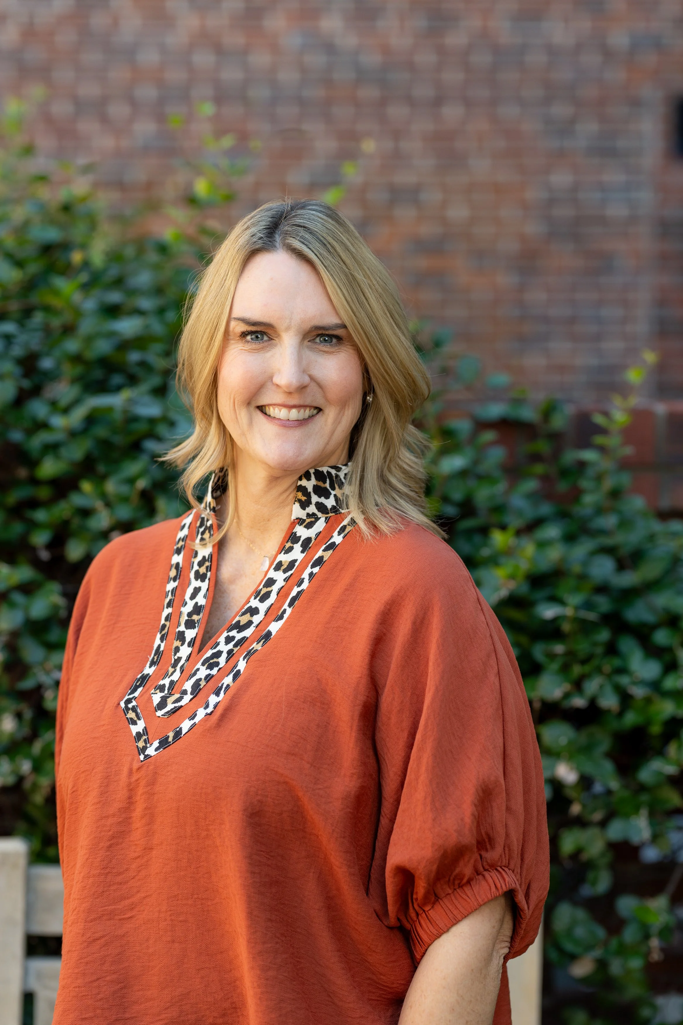 Woman with blonde hair smiling outdoors, wearing an orange top with leopard print accents, standing in front of greenery and a brick wall.