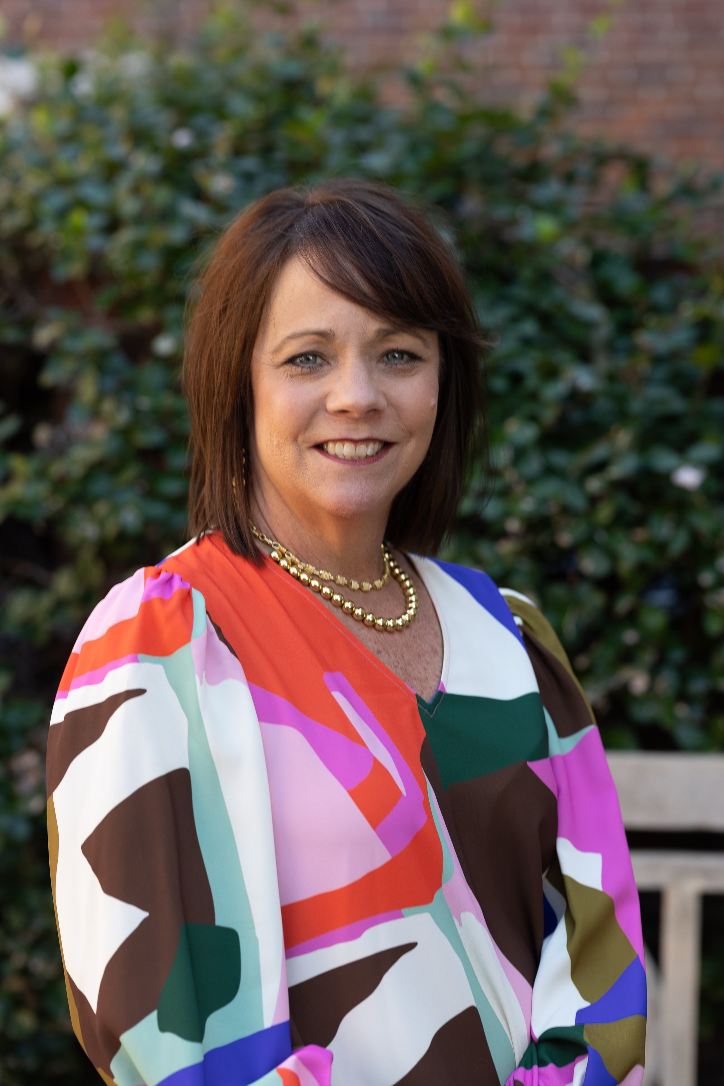 A woman with shoulder-length brown hair wearing a colorful, patterned blouse and a gold necklace, standing outdoors in front of a leafy green background.