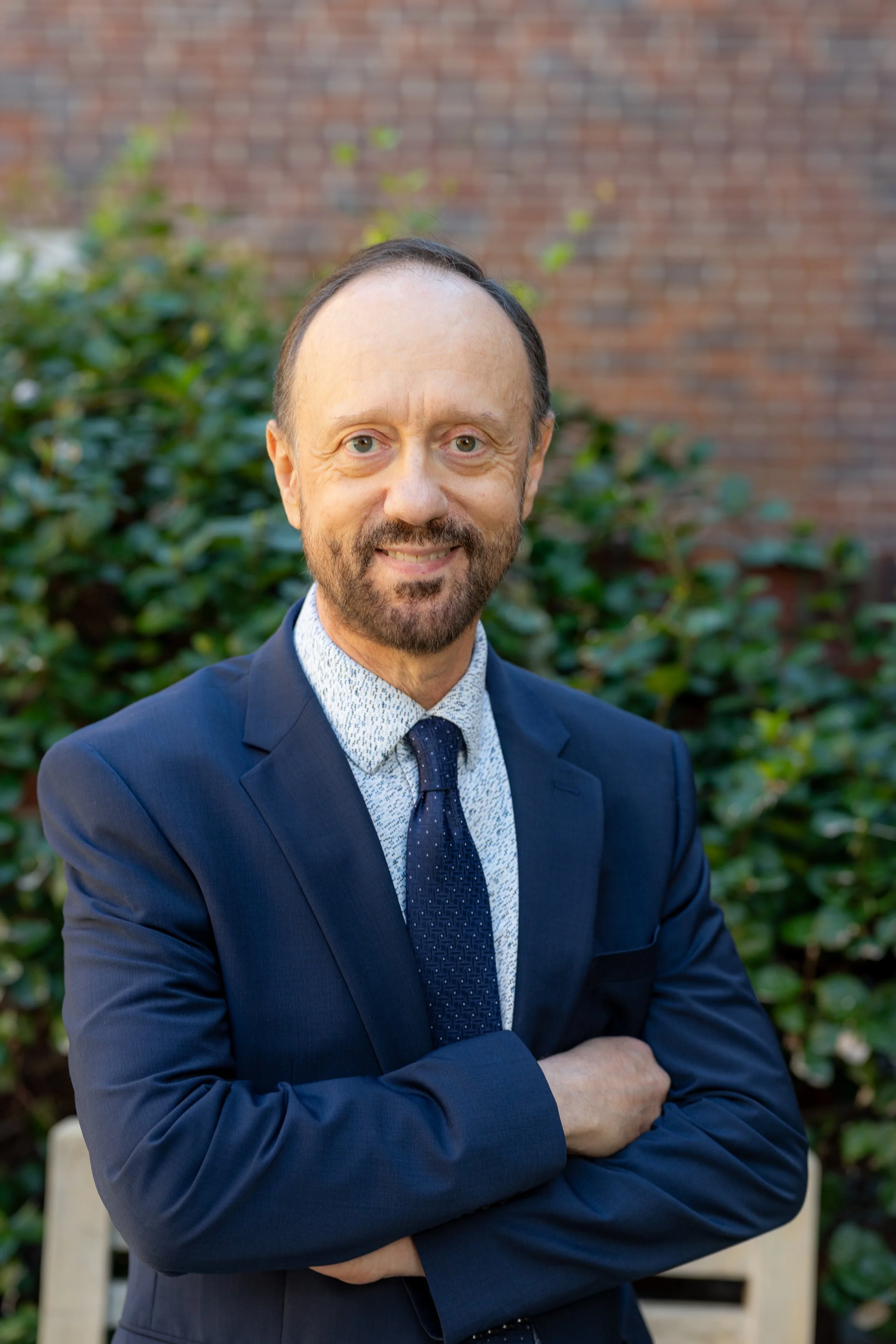 A man with light skin, a beard, and a receding hairline smiling outdoors in front of green foliage and a brick wall. He is wearing a dark suit, a light-colored dotted shirt, and a dark tie.