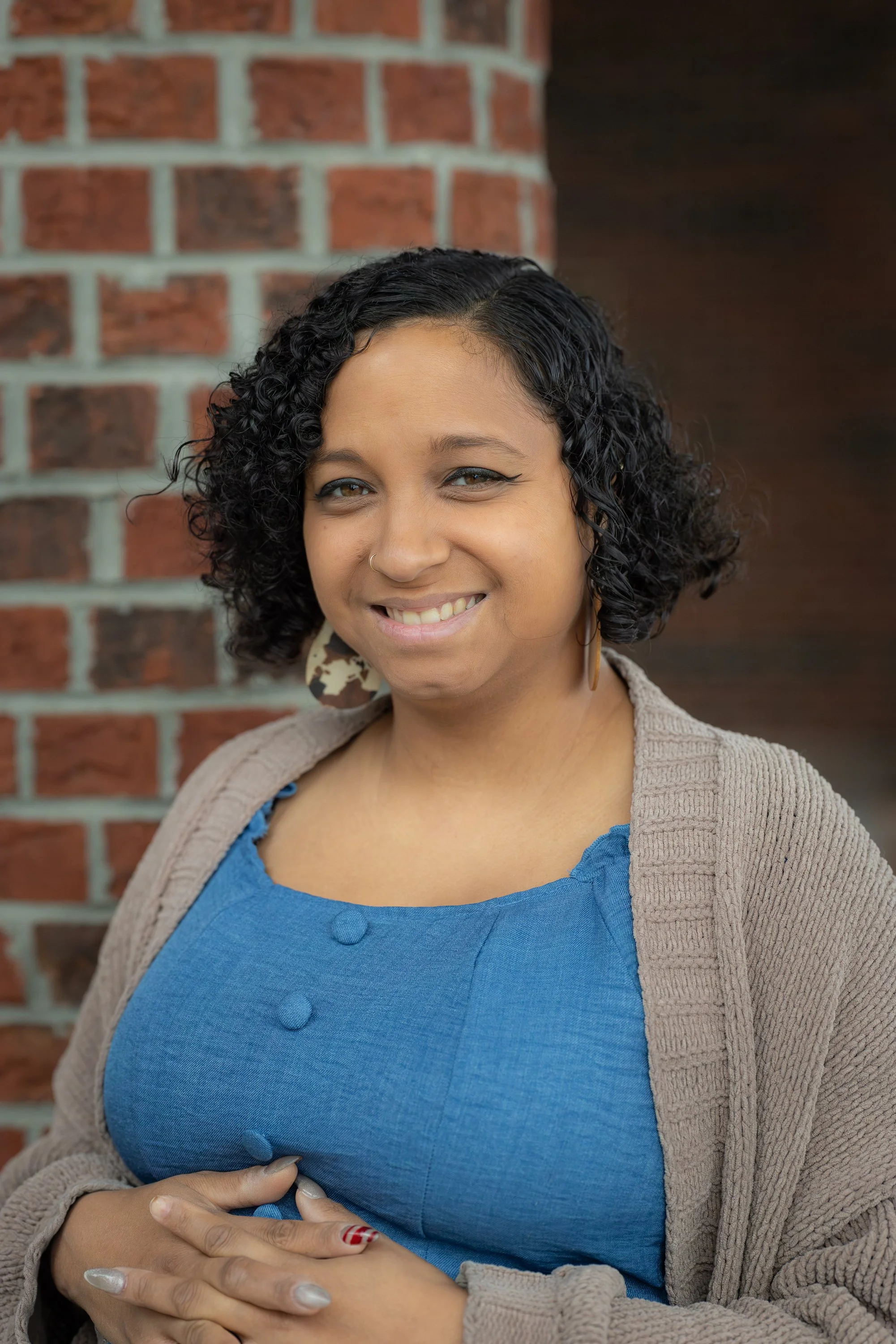 Smiling woman with curly black hair, wearing a blue top, beige cardigan, and large earrings, standing in front of a brick wall.