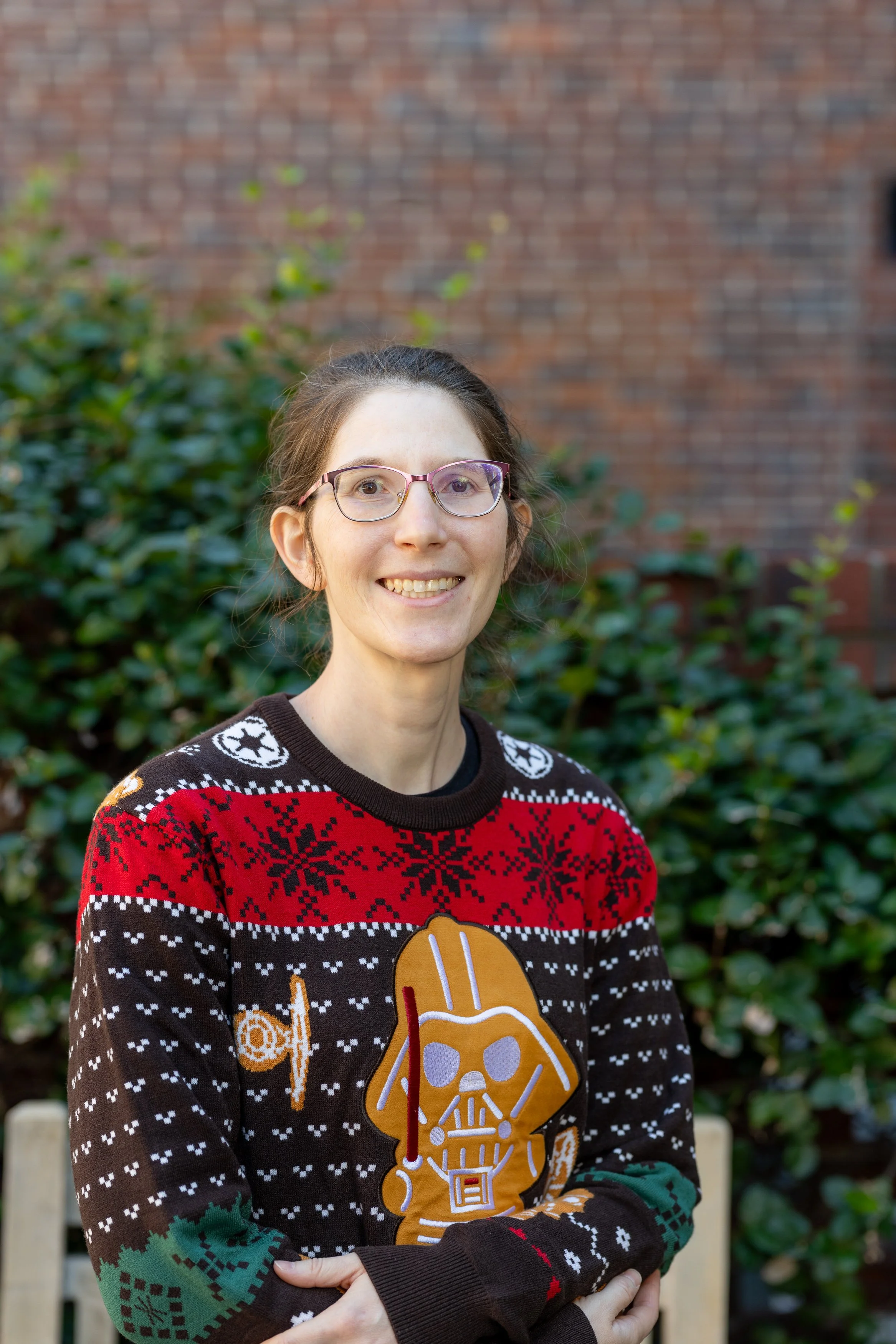 A woman wearing glasses and a Christmas sweater with Star Wars characters and holiday patterns, standing outdoors in front of green bushes and a brick wall.