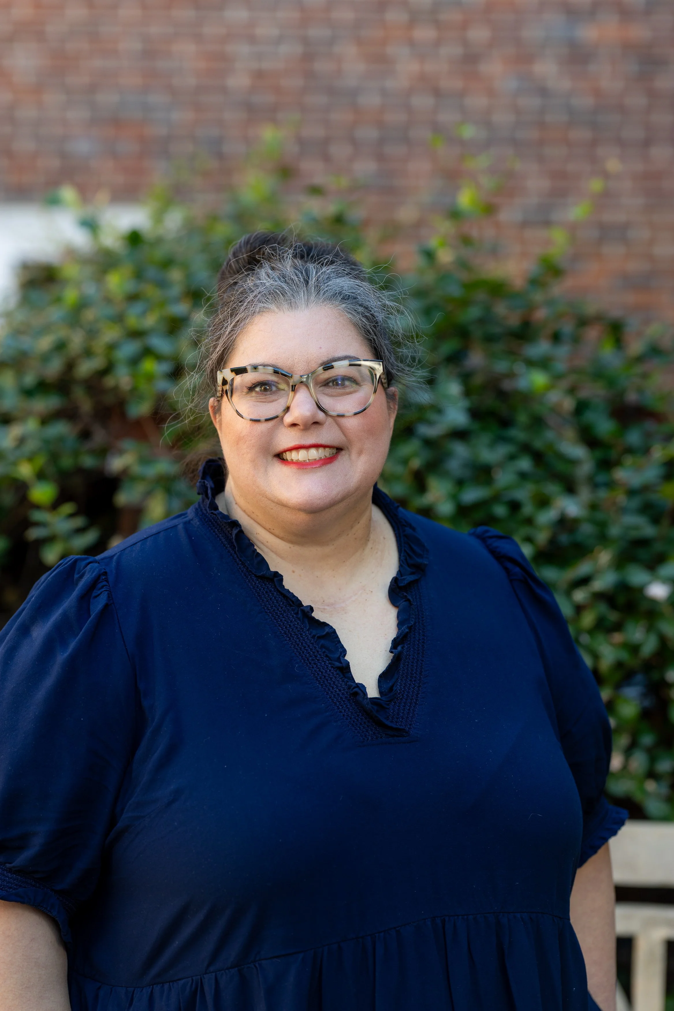A smiling woman with glasses and dark hair in a ponytail, wearing a dark blue top, standing outdoors in front of green bushes and a brick wall.