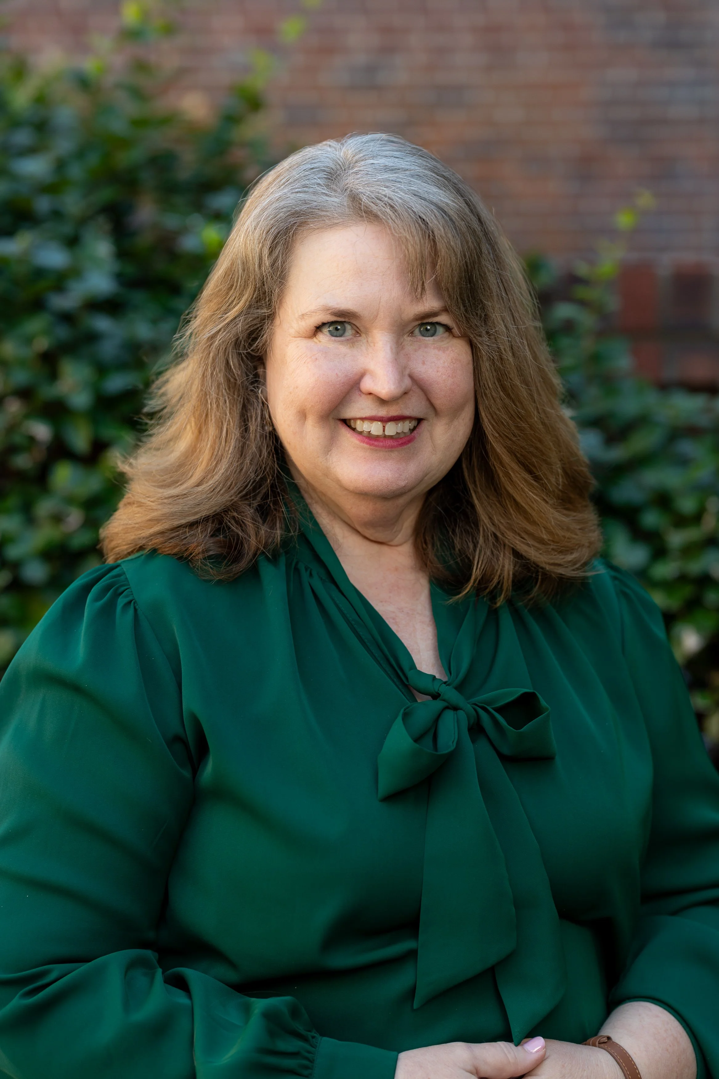 A woman with shoulder-length reddish-brown hair smiling inside a church. She is wearing a black blazer, a white shirt, and a necklace with a cross pendant. Behind her is a colorful stained glass window and wooden pews in the church interior.