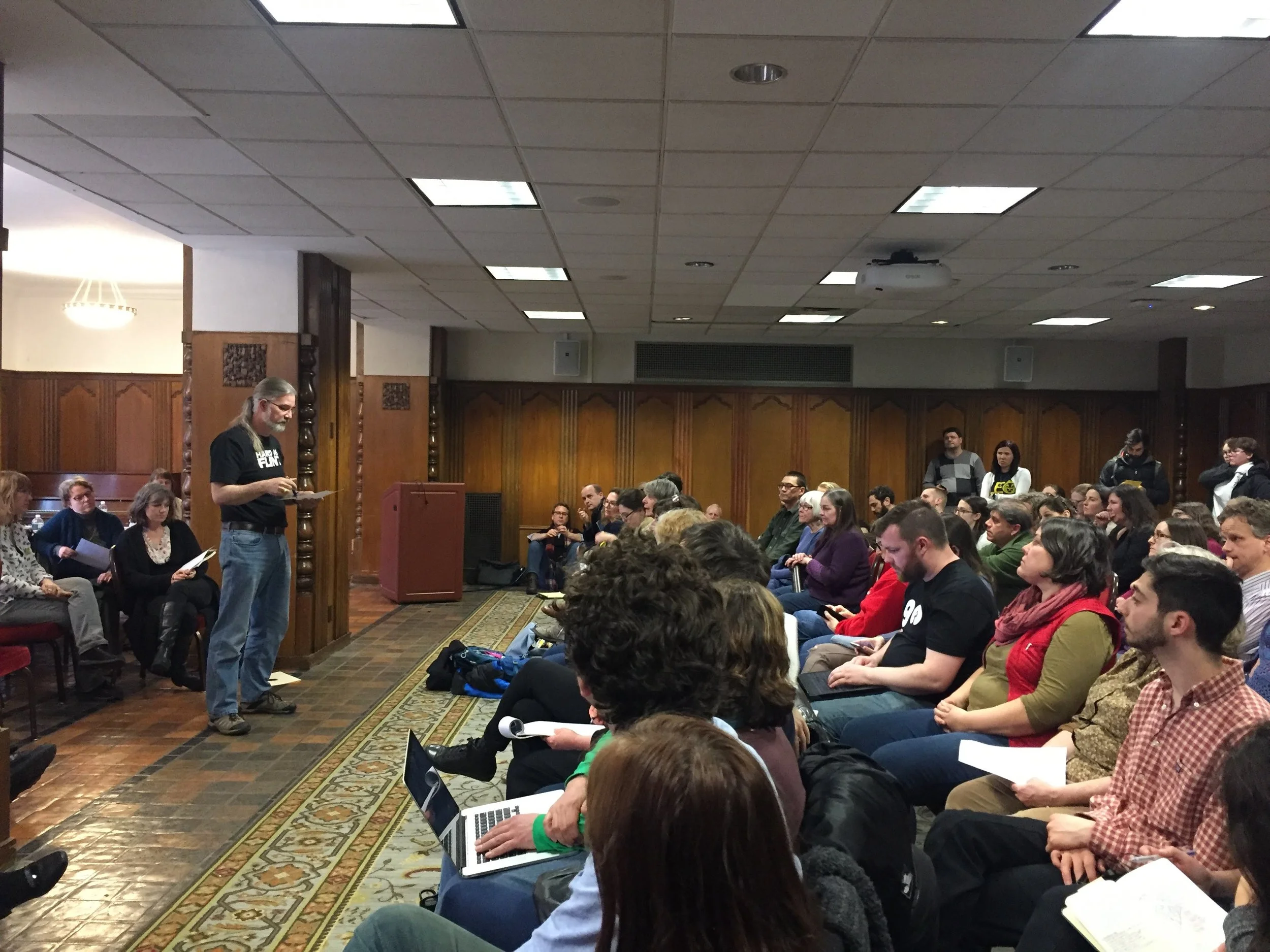 A packed room--on the right, many rows of chairs, all filled, people along the walls on the sides and in back. On the left, bargaining team member Stevens Wandmacher provides an update.