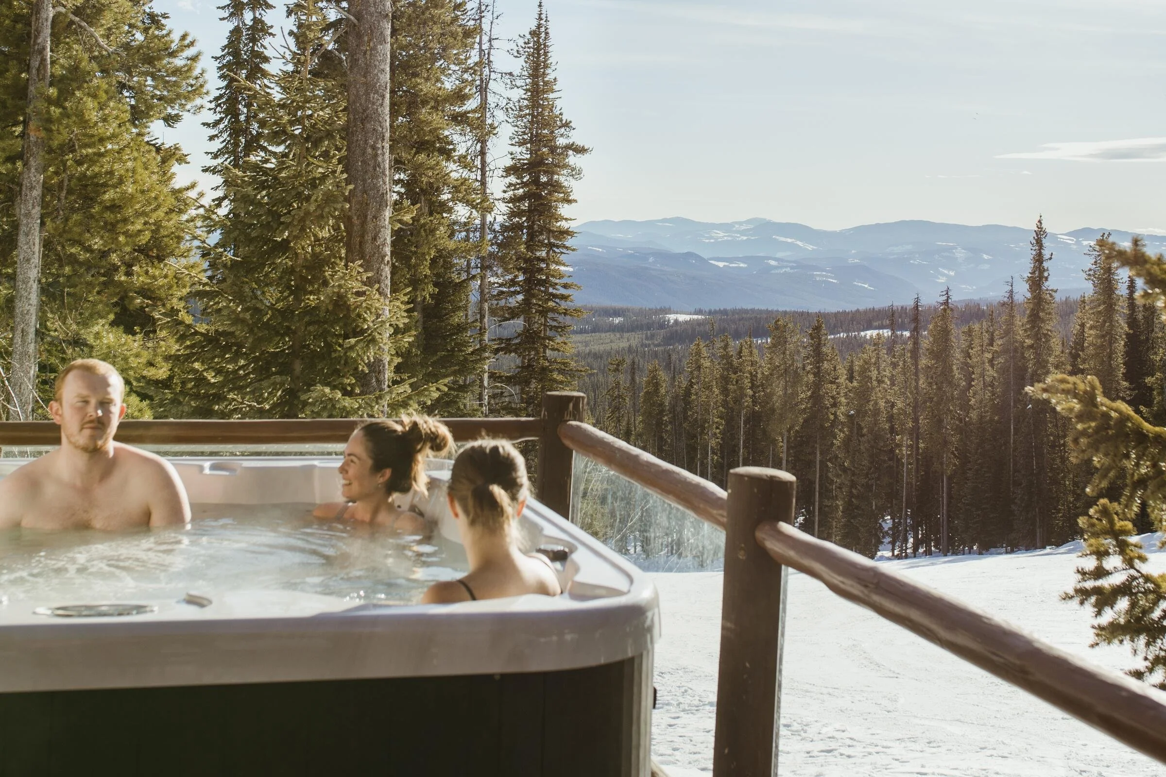 Three people in a hot tub on the deck of a Big White accommodation talking and smiling with snow-capped mountains in the back ground.