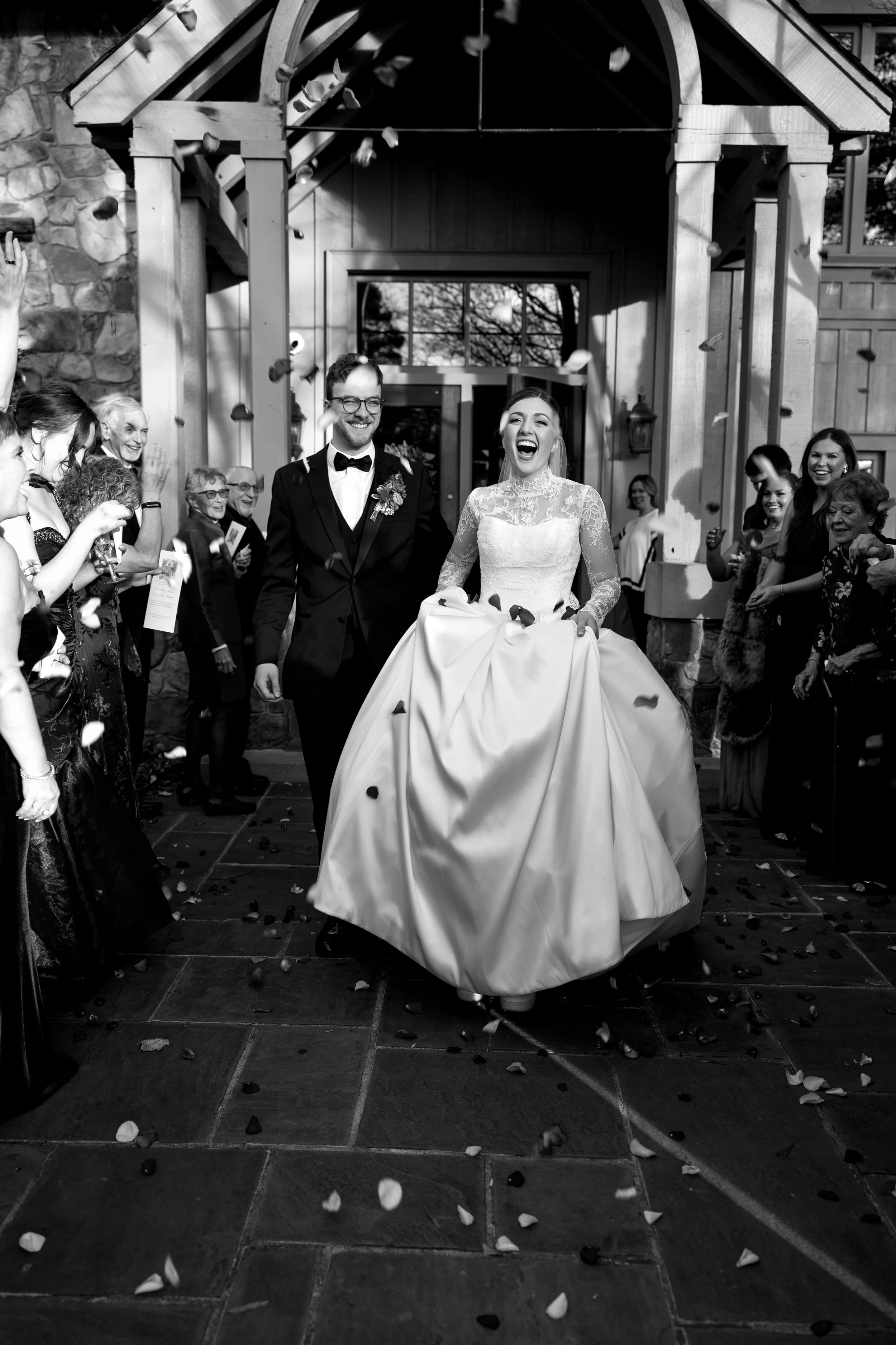 Black and white photo of a newlywed couple exiting a building, surrounded by friends and family throwing confetti, with the bride smiling and holding up her wedding dress.