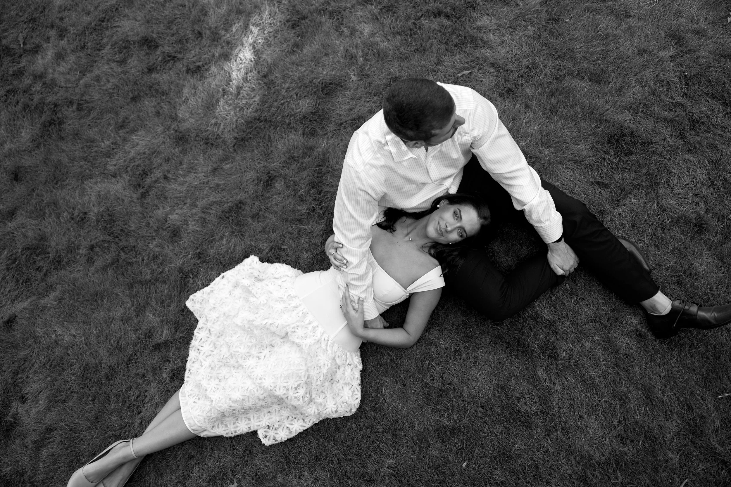 A man and woman lie on grass, with the woman resting her head on the man's lap. The woman wears a white dress and gazes upward, while the man looks down at her, holding her hand.