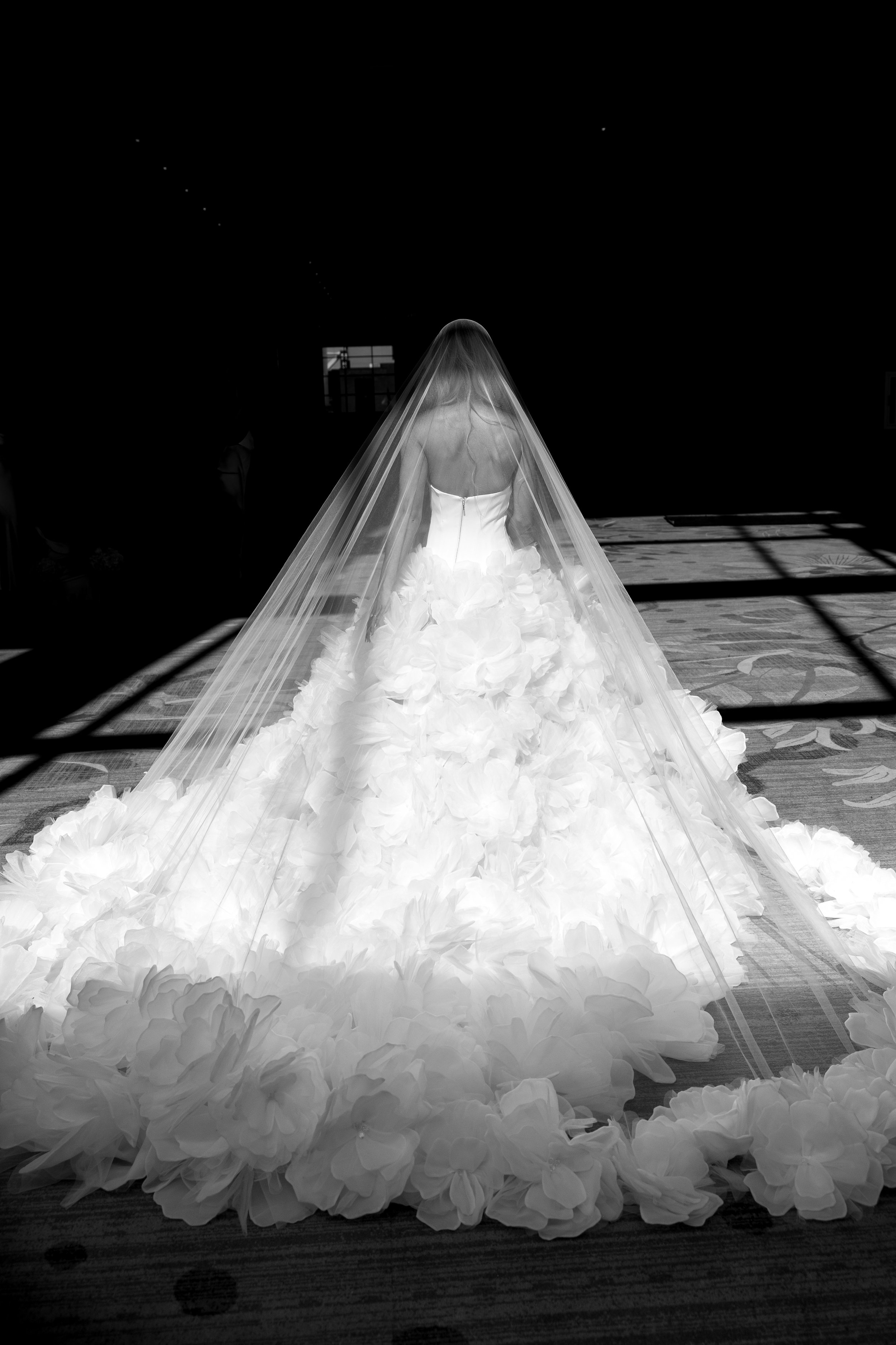 A bride in a white wedding gown with a long train adorned with large fabric flowers, wearing a veil, standing indoors with sunlight casting shadows.