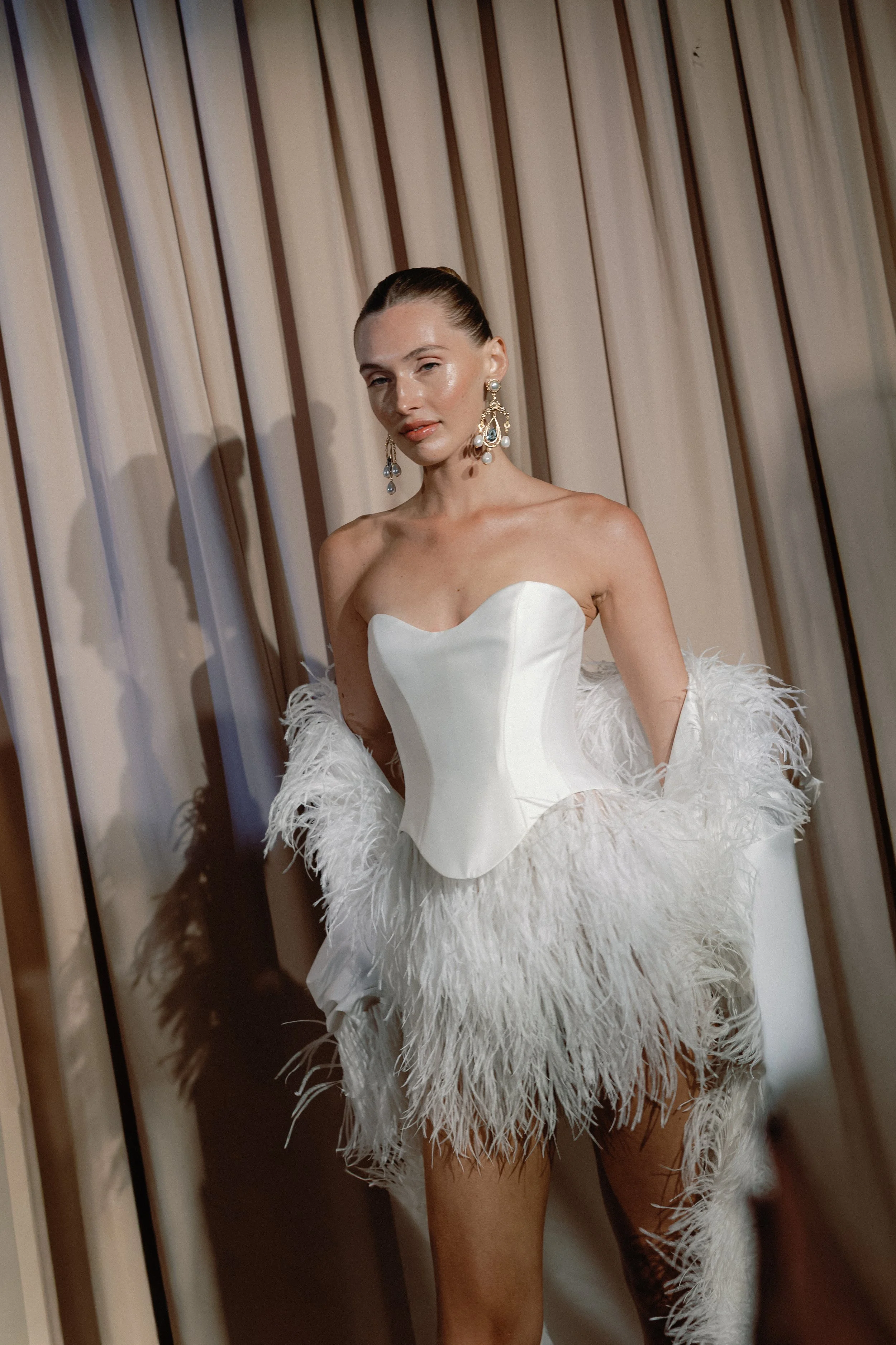 A woman wearing a white strapless dress with feathered jacket standing in front of a beige curtain.